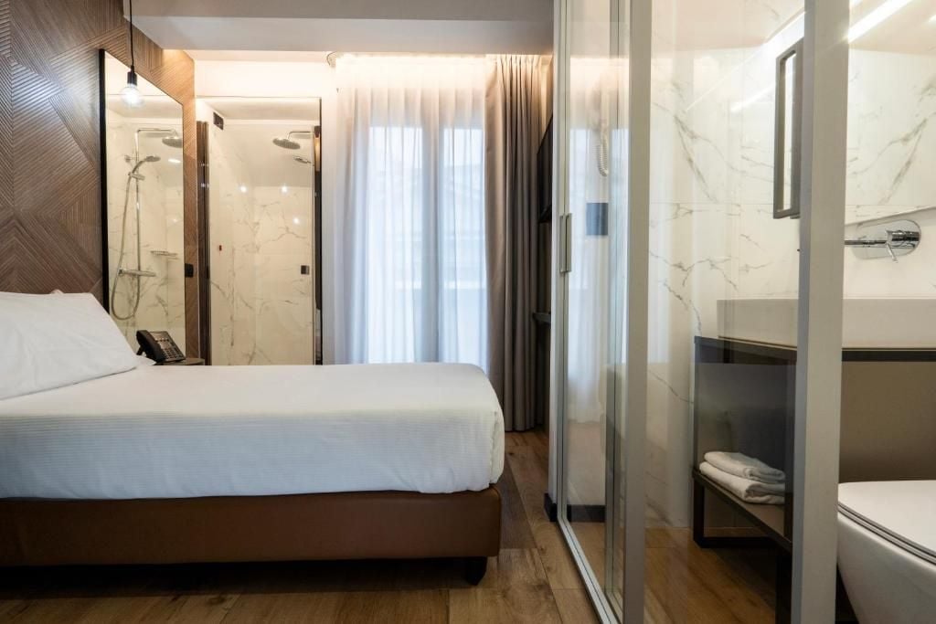 Una camera d'albergo con un letto, un box doccia in vetro, una toeletta con specchio e una finestra con tende.