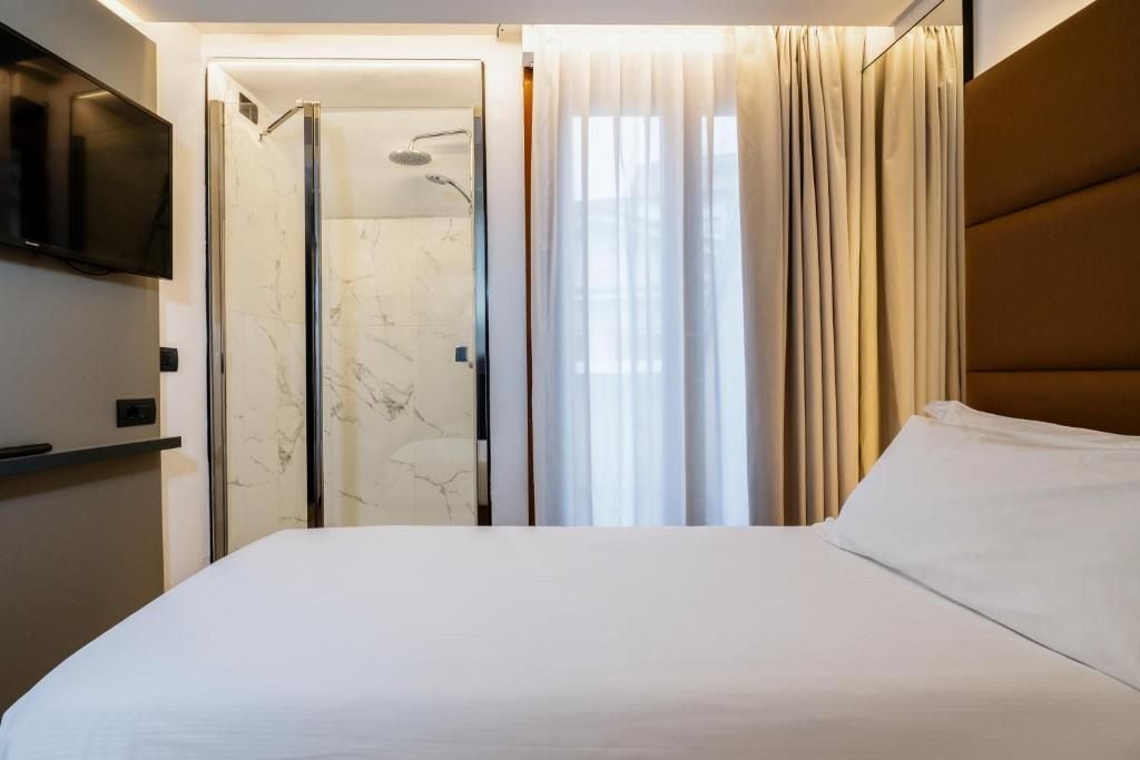 A hotel room with a bed, a television mounted on the wall, a shower area with a glass door, and a window with curtains.