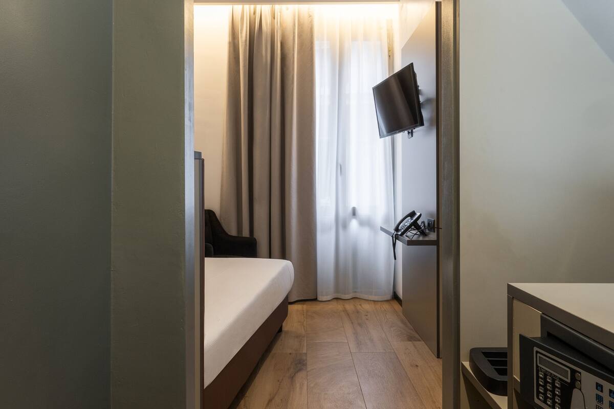 Una moderna camera d'albergo con pavimento in legno, un letto, una scrivania con un computer, una TV a parete e una finestra con tende.