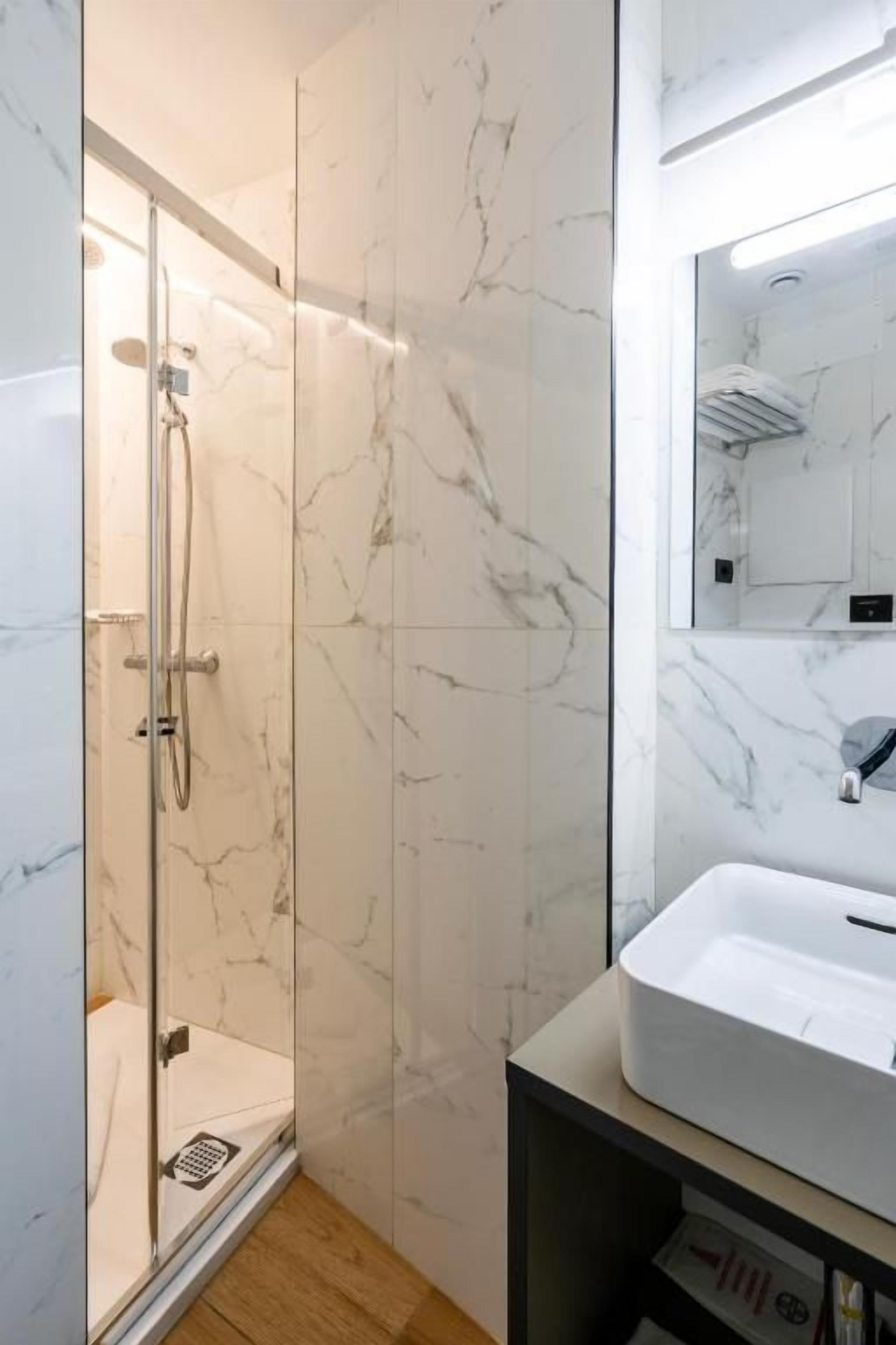 Un baño moderno con ducha envuelta en vidrio, un lavabo blanco y paredes de mármol.