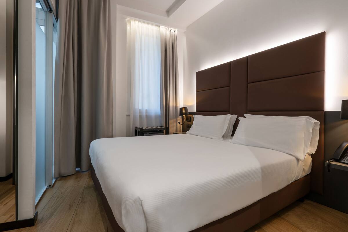 Una moderna camera d'albergo con un letto grande, un comodino, una lampada e vista all'esterno attraverso una finestra con tende.