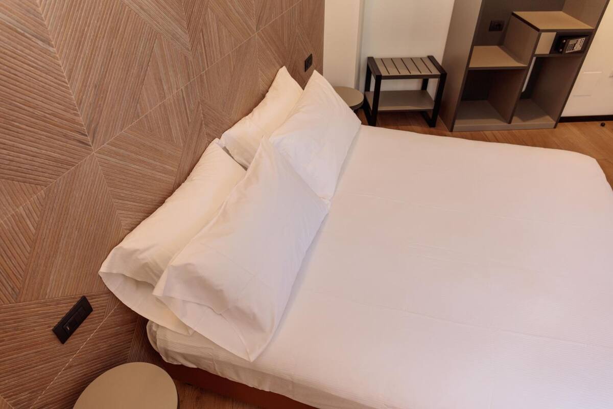 Un letto rifatto con lenzuola bianche in una camera moderna, con una testiera a motivi a spina di pesce e un piccolo comodino in legno.