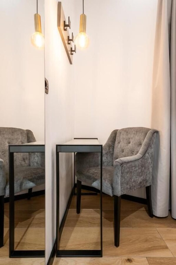A room with a grey upholstered chair, a small table with a black frame, and a mirror reflecting the room. There are two light fixtures on the wall. - London Hotel (Milan)