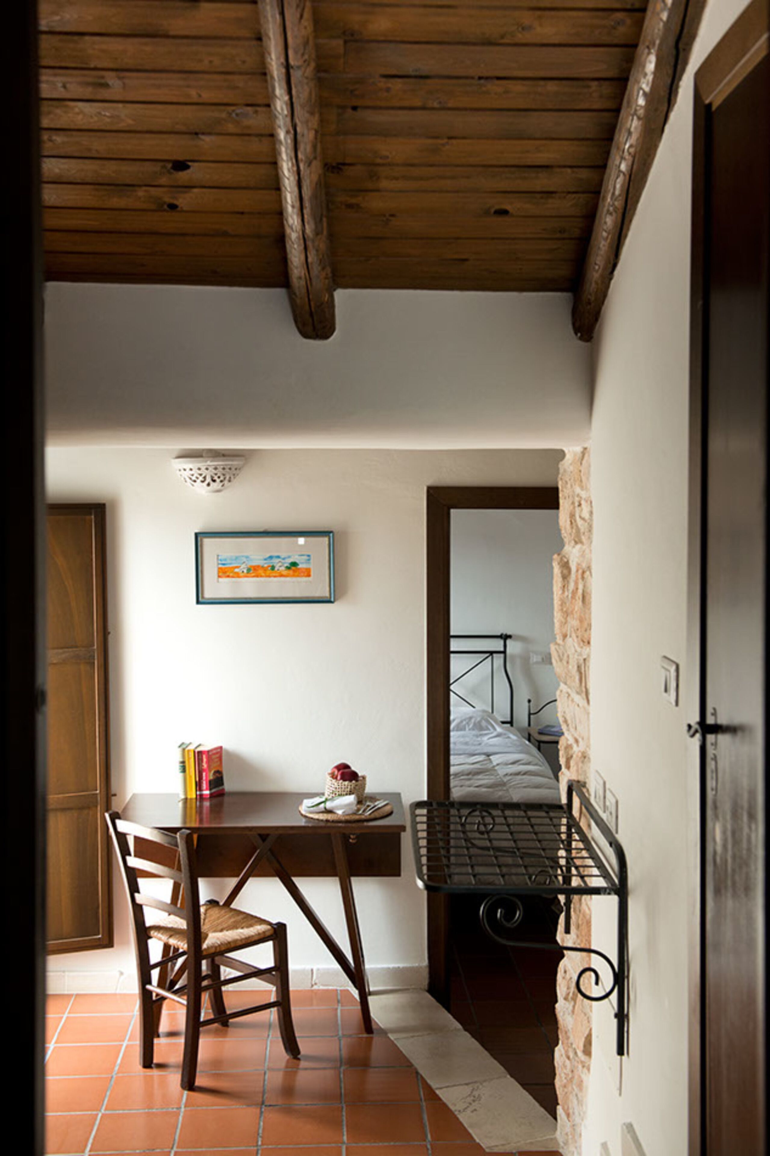 A small room with a wooden ceiling, a bed, a desk with a chair, and a framed picture on the wall.