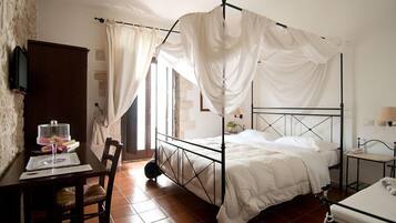 Ein Bett mit Baldachin, ausgestattet mit weißer Bettwäsche, ein Holztisch mit Stuhl, ein kleiner Tisch mit Lampe und ein Fenster mit Vorhängen.