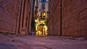 Una estrecha calle adoquinada flanqueada por edificios de piedra, que conduce a un edificio bien iluminado con un balcón.
