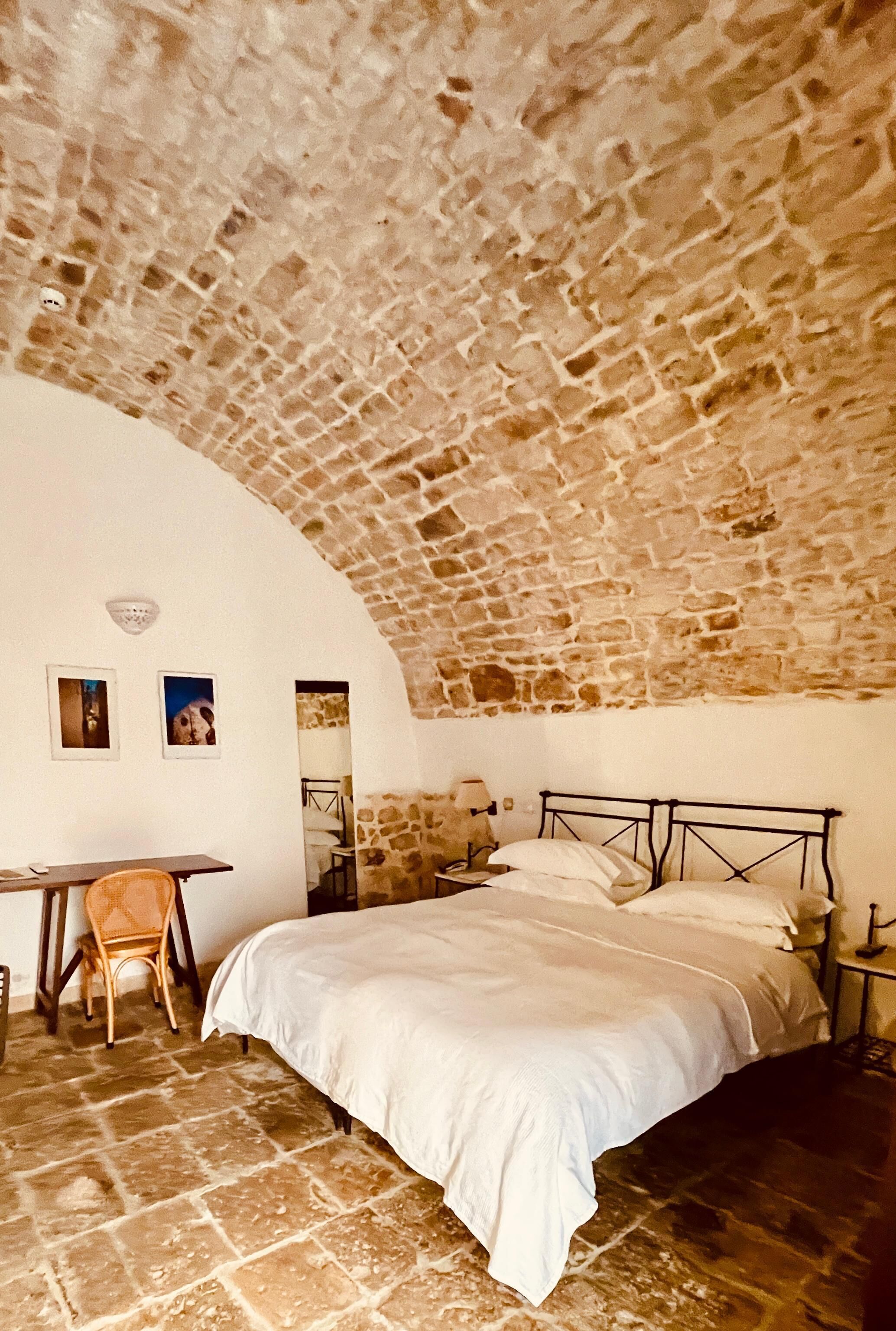 A room with a stone ceiling, a bed with a metal frame, a chair, and a table.