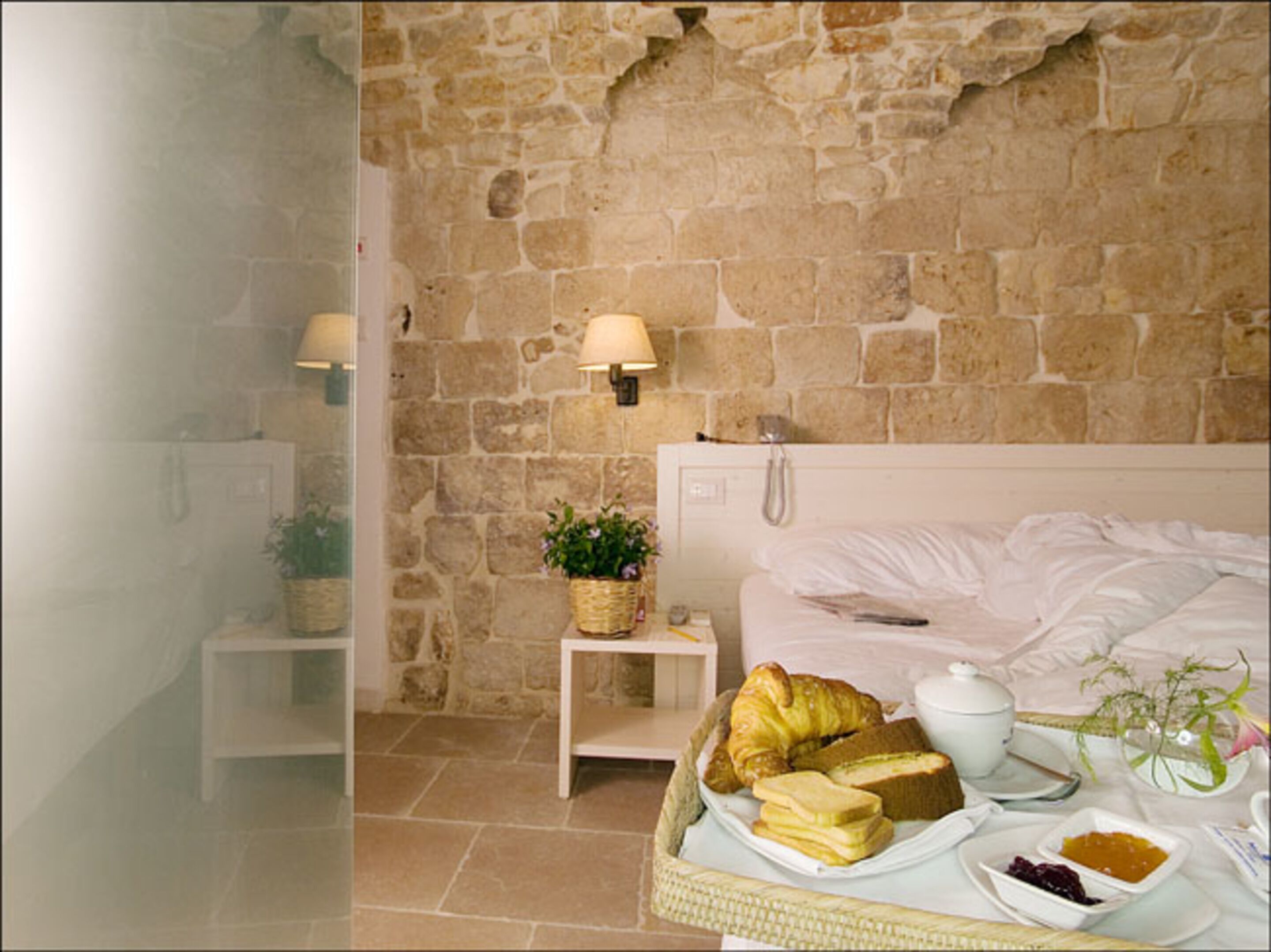 A room with a stone wall, a bed with white linens, a bedside table with a lamp, a small plant, and a basket of food.