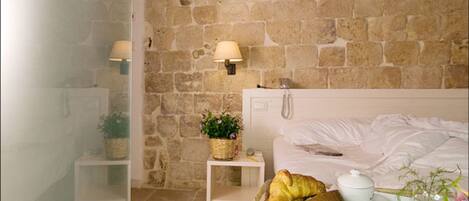 A room with a stone wall, a bed with white linens, a bedside table with a lamp, a small plant, and a basket of food.