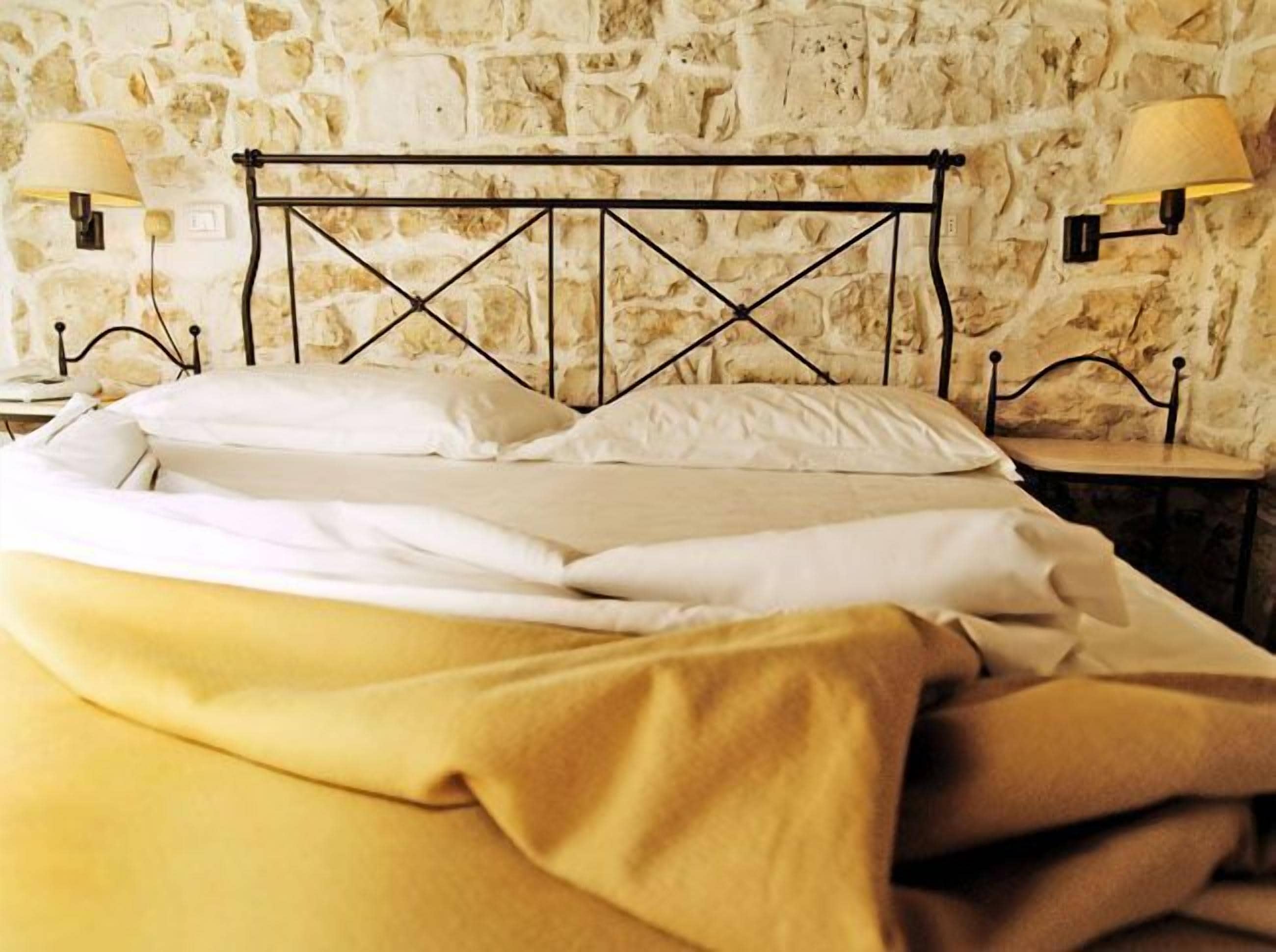 Un lit avec une tête de lit en métal, une literie blanche et une couverture jaune. Deux lampes murales sont fixées de chaque côté du lit.