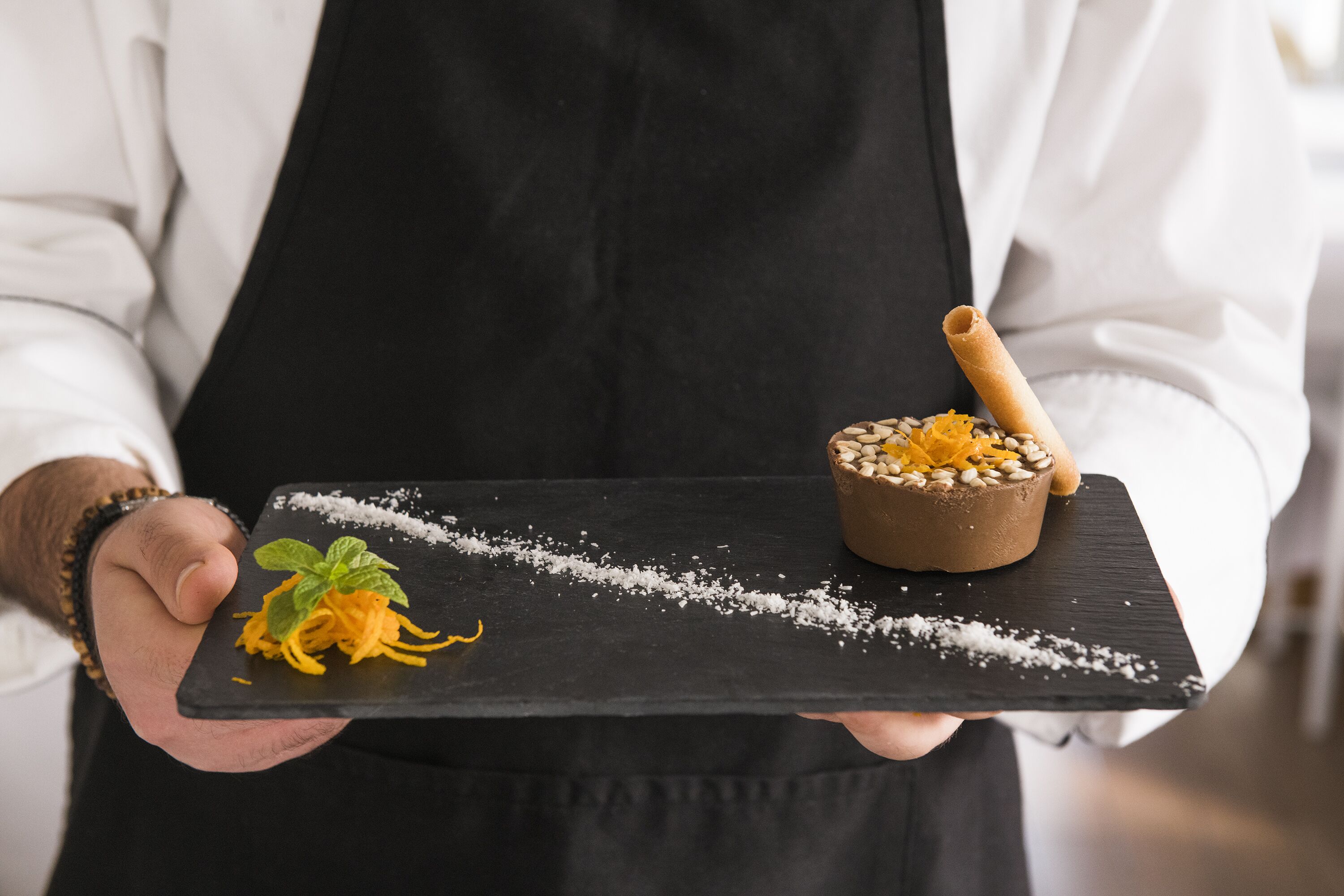 Una persona sostiene una tabla de pizarra negra con un postre y un tazón pequeño de nueces.