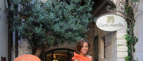 A woman in an orange dress standing next to a white bicycle with baskets, in a narrow street with stone buildings and greenery.