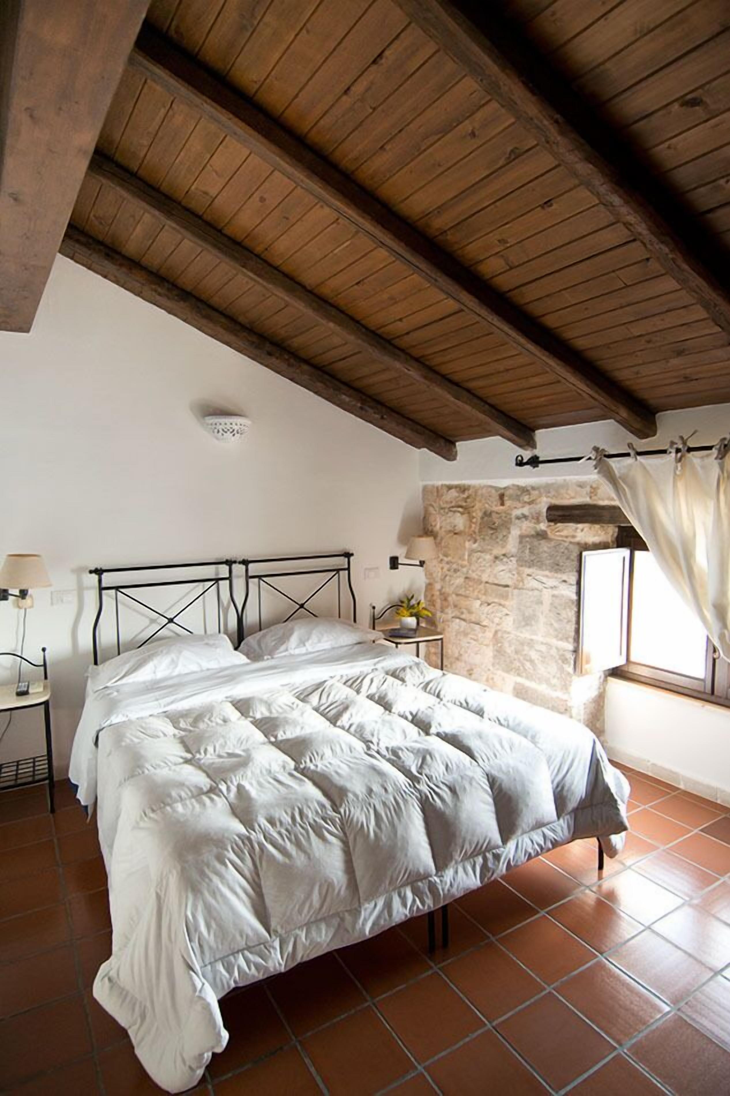 A bedroom with a bed, a stone wall, a window with curtains, and a wooden ceiling.
