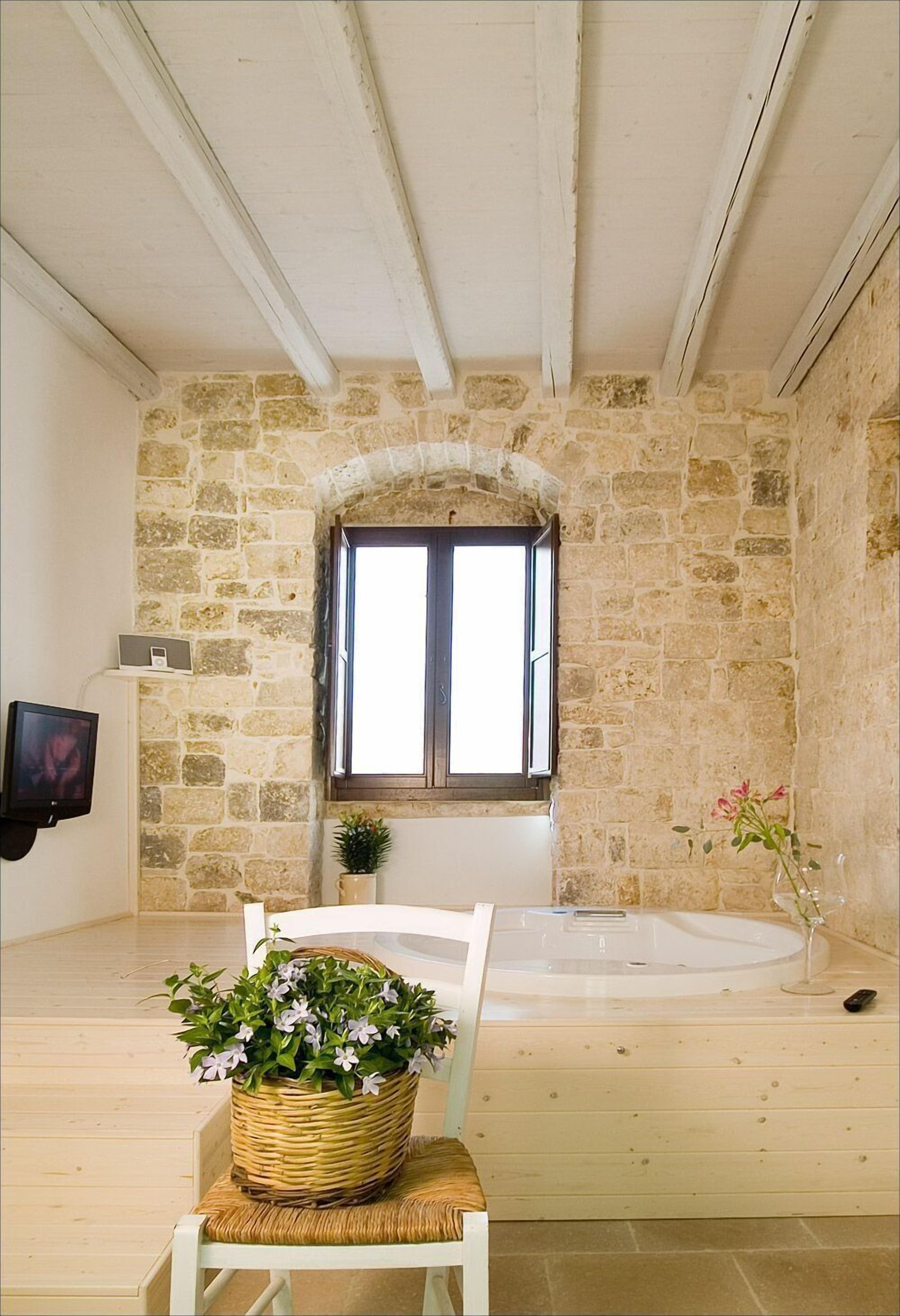 A room with stone walls, a wooden floor, a white chair, a wicker basket with flowers, a white bathtub, and a window with a view.