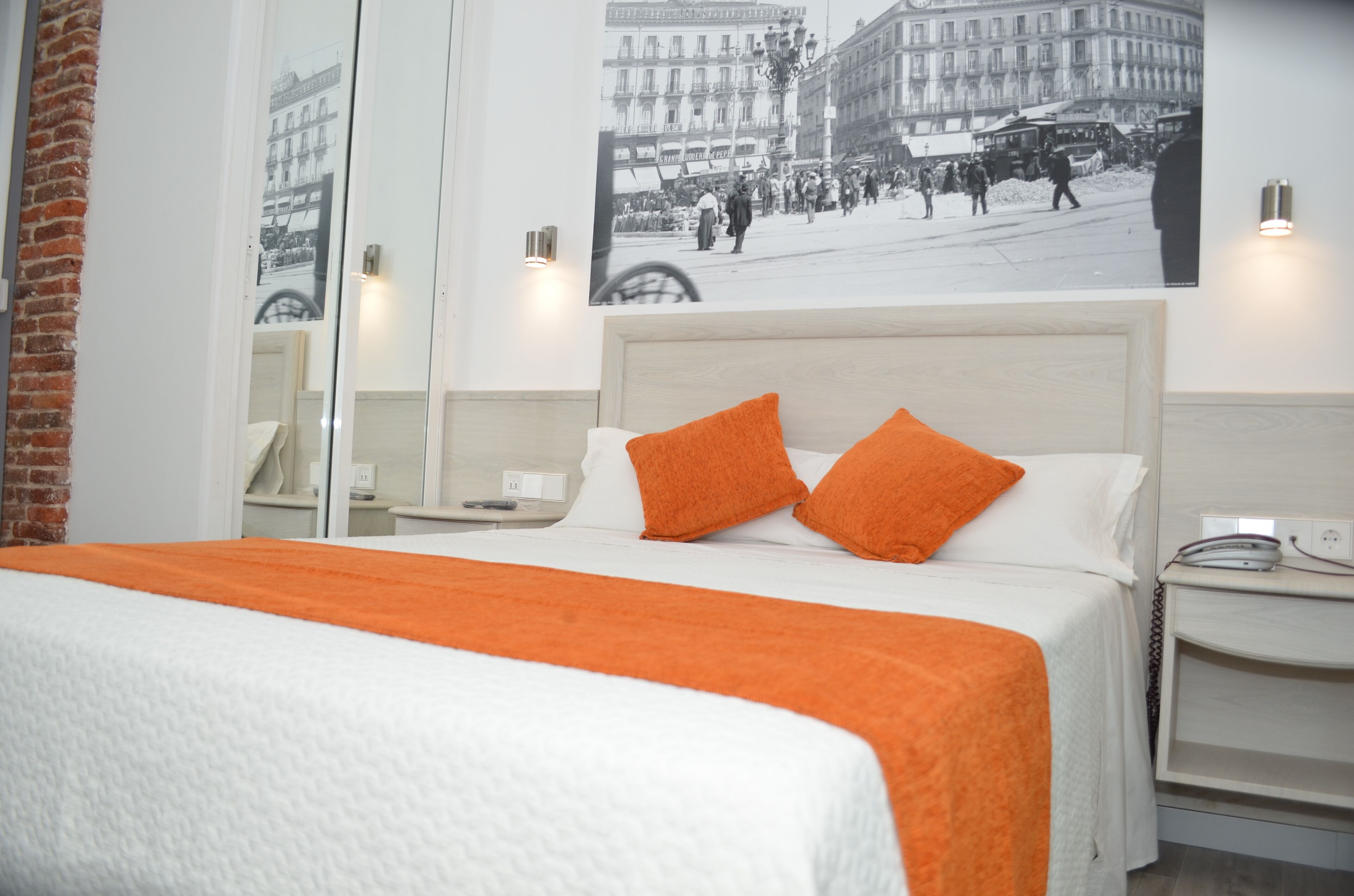 A hotel room with a bed, two orange pillows, a nightstand, a lamp, and a wall-mounted picture of a city scene.