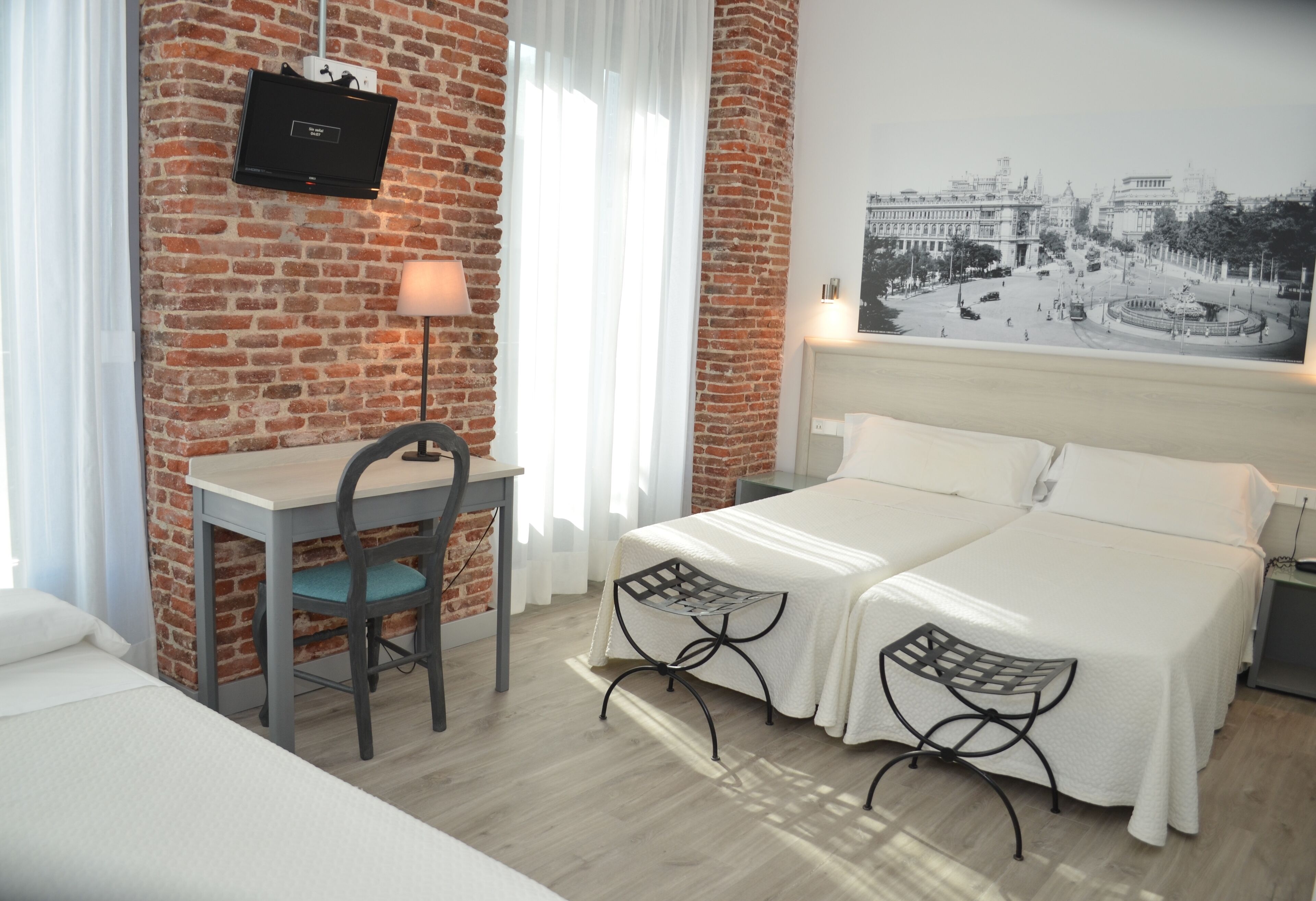 A hotel room with a brick wall, a desk with a chair, two beds, and a large framed picture on the wall.
