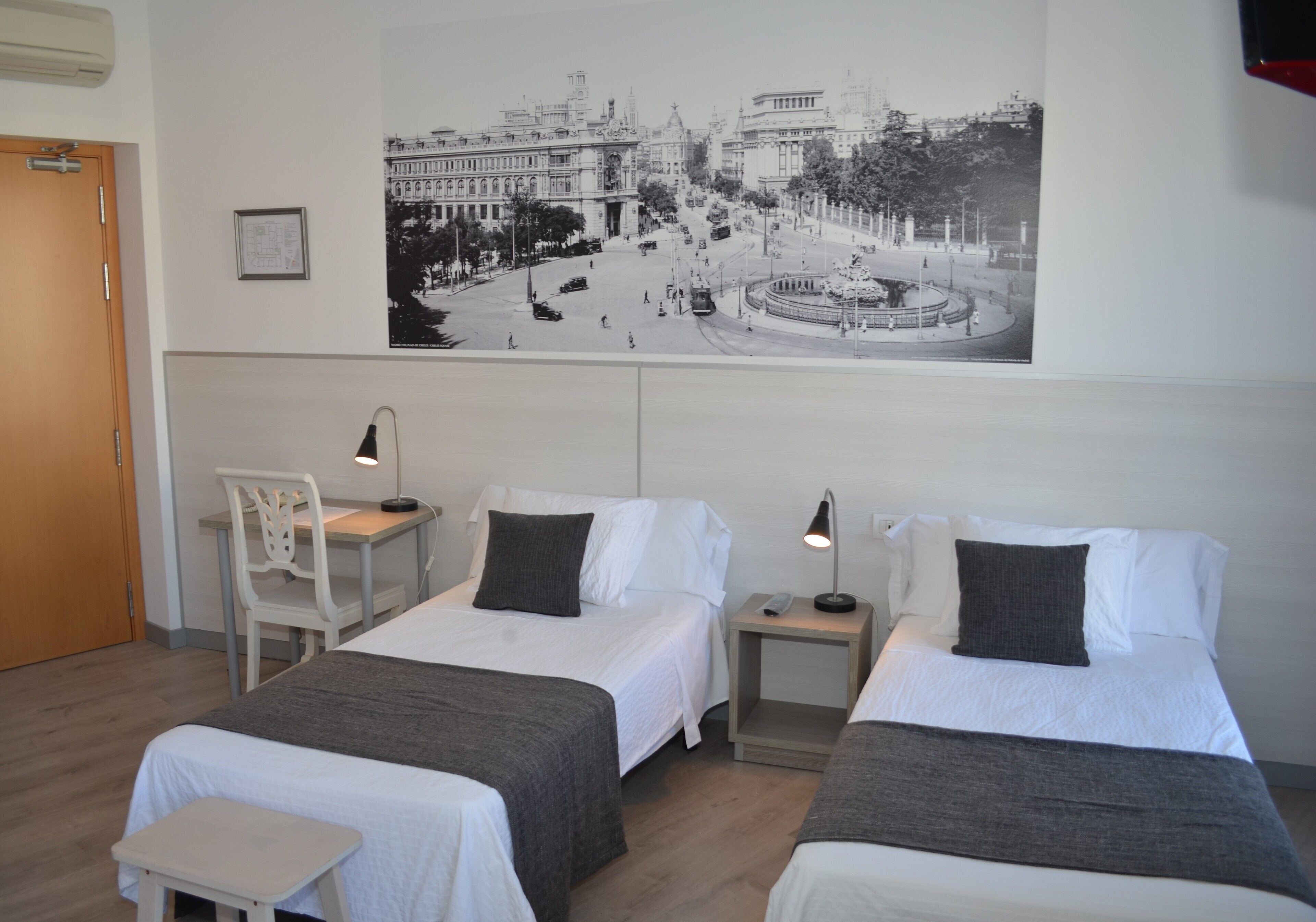 A hotel room with two beds, a desk, a chair, and a large cityscape photograph on the wall.