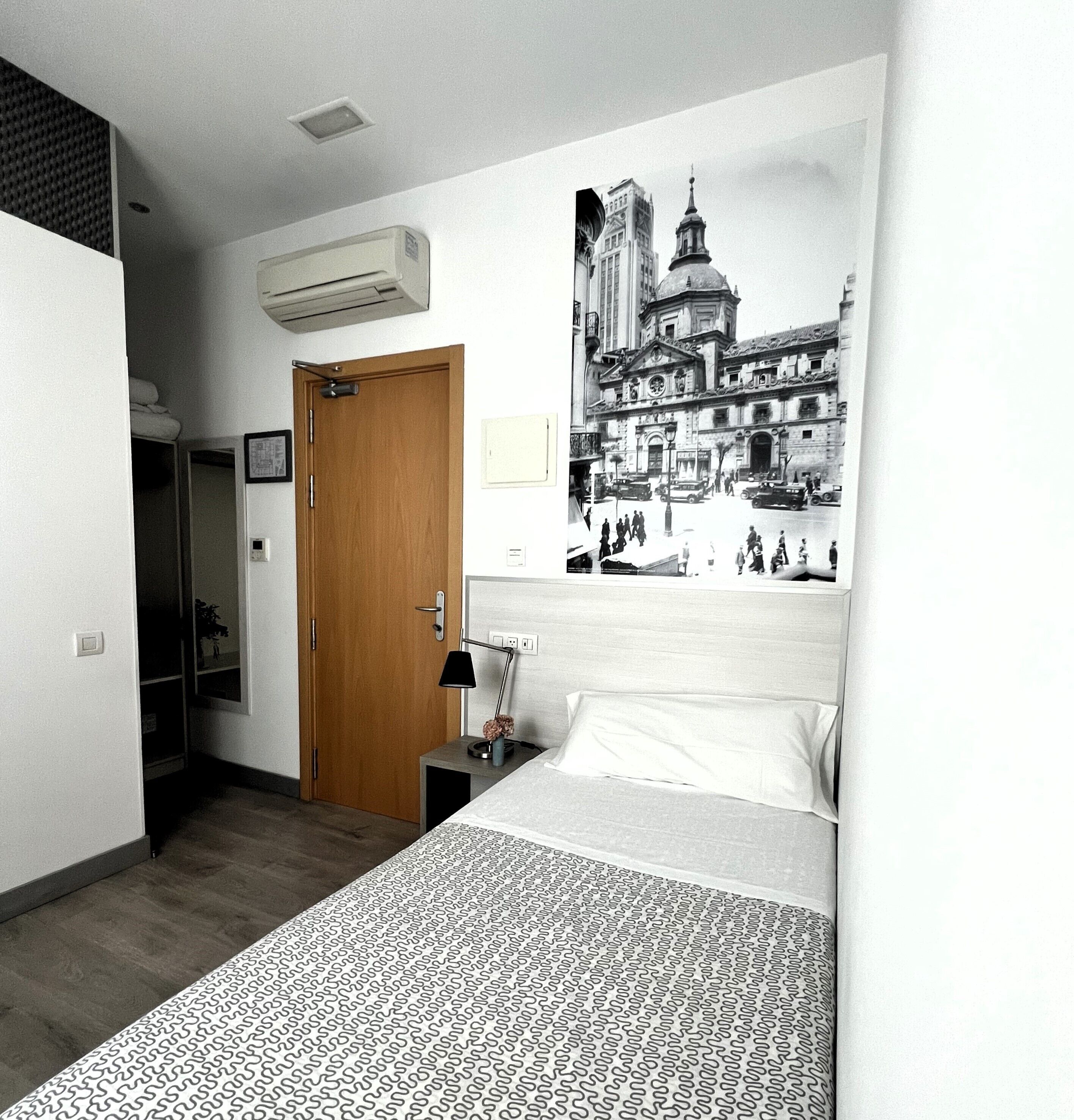 Una habitación de hotel con una cama, una mesita de noche, una lámpara, un equipo de aire acondicionado, una puerta e imagen en la pared que muestra un paisaje urbano.