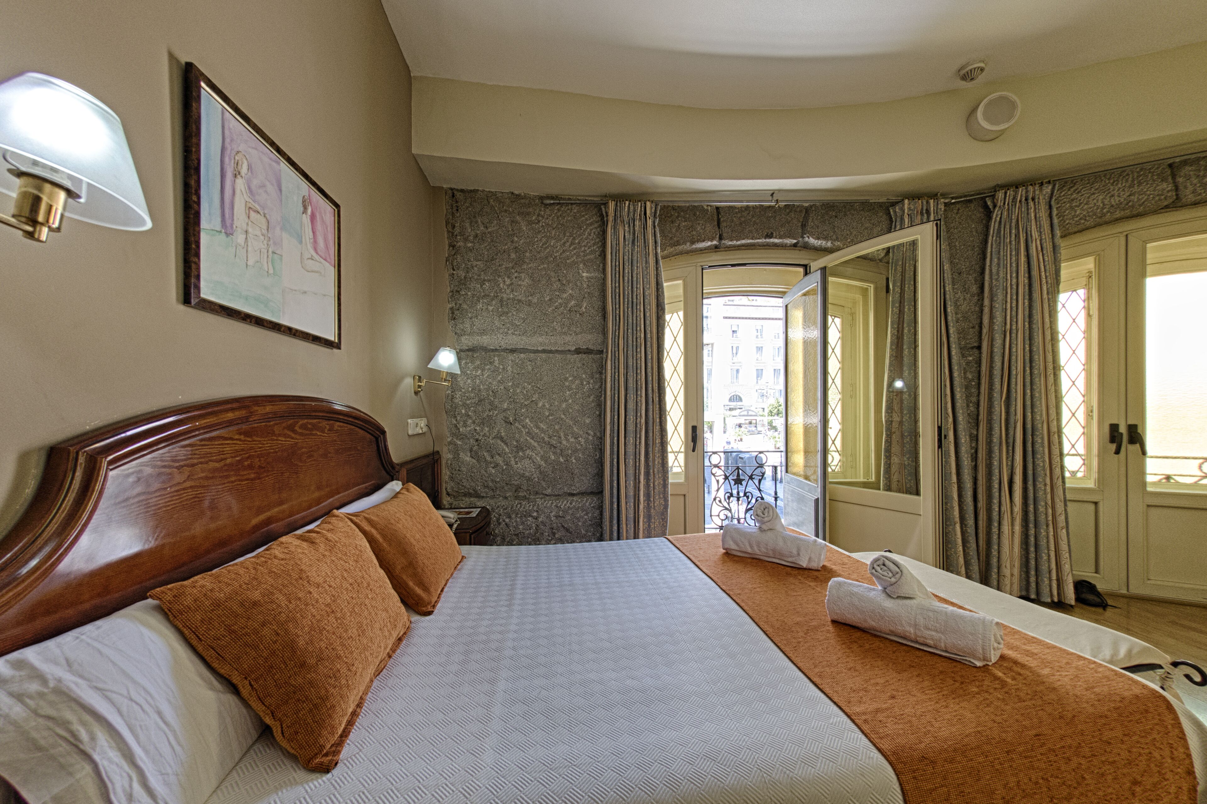 Een hotelkamer met een groot bed, een houten hoofdbord, een nachtkastje, een lamp, een ingelijst foto aan de muur en uitzicht op het balkon door een deur.
