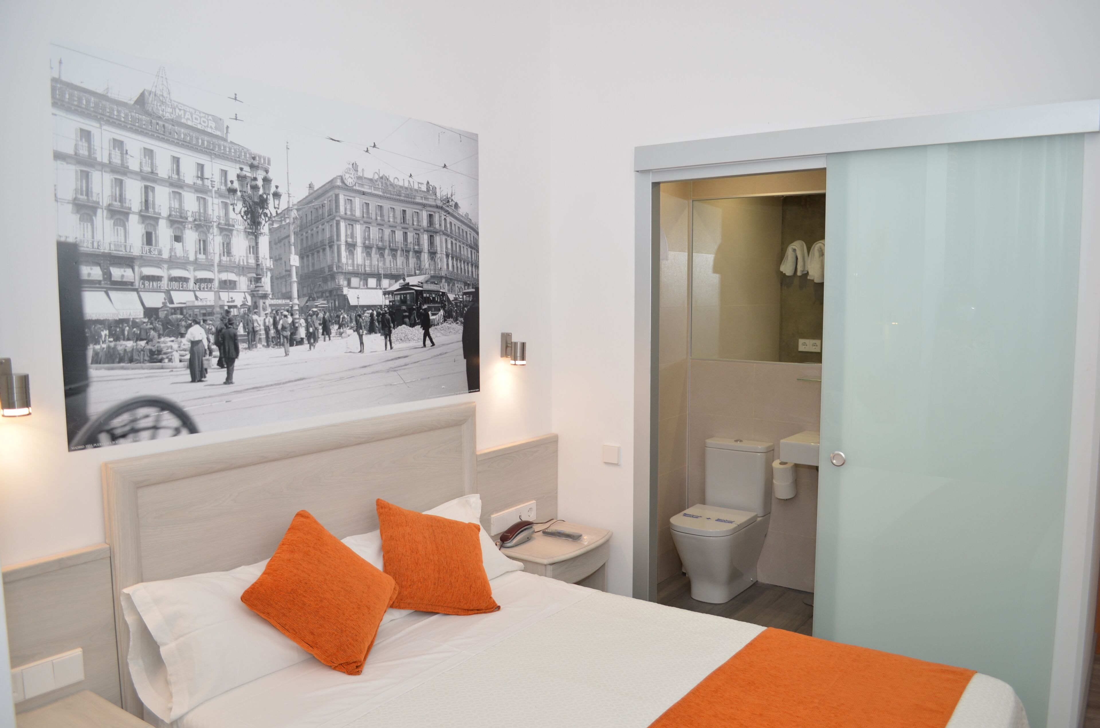 A hotel room with a bed, two orange pillows, a nightstand, a wall-mounted lamp, a bathroom with a toilet and sink, and a poster of a street scene.