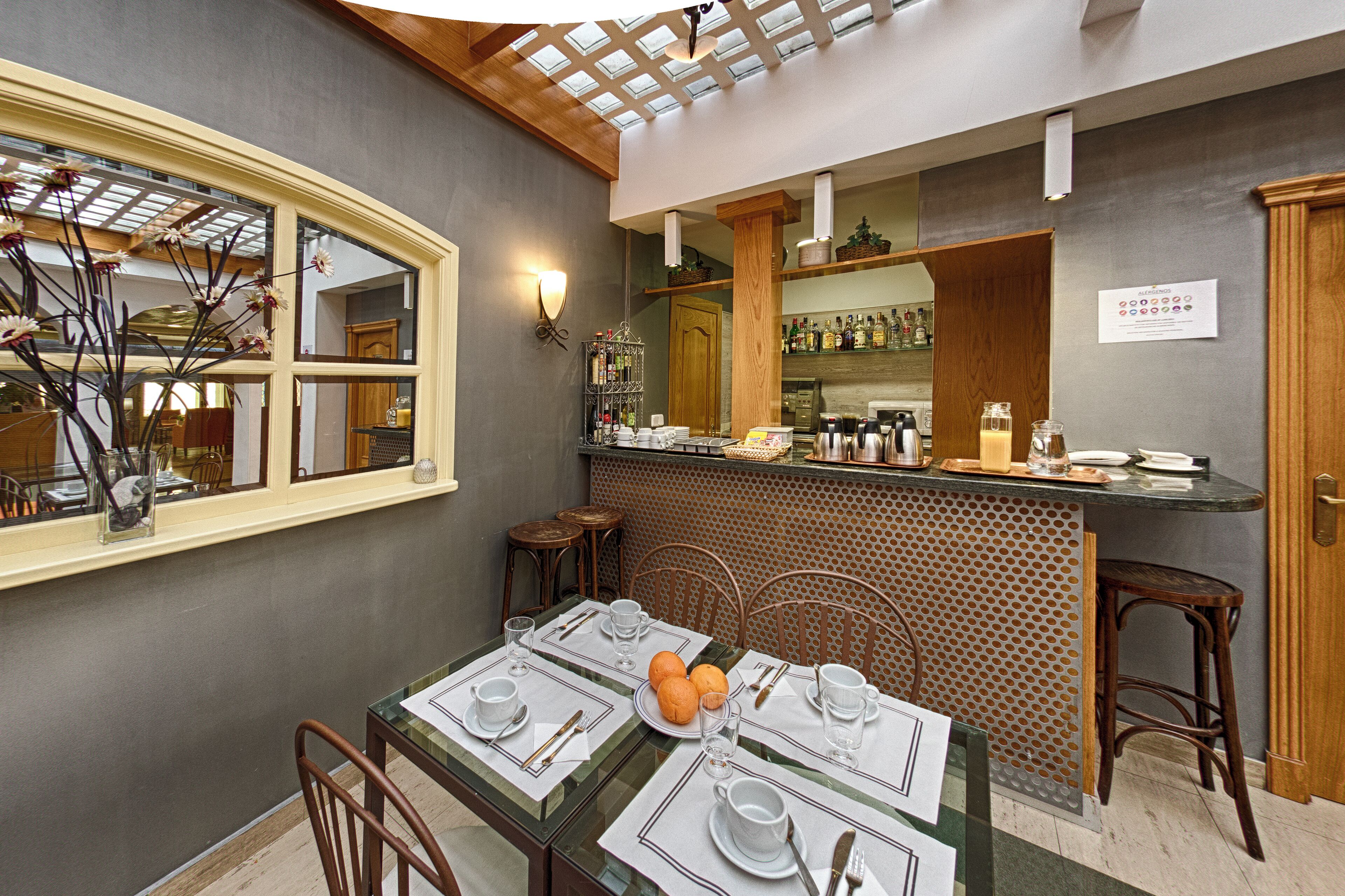 Un coin repas avec une table en verre dressée pour quatre personnes, un bar doté d’un comptoir en bois et une vue sur la cuisine par le biais d’une fenêtre.