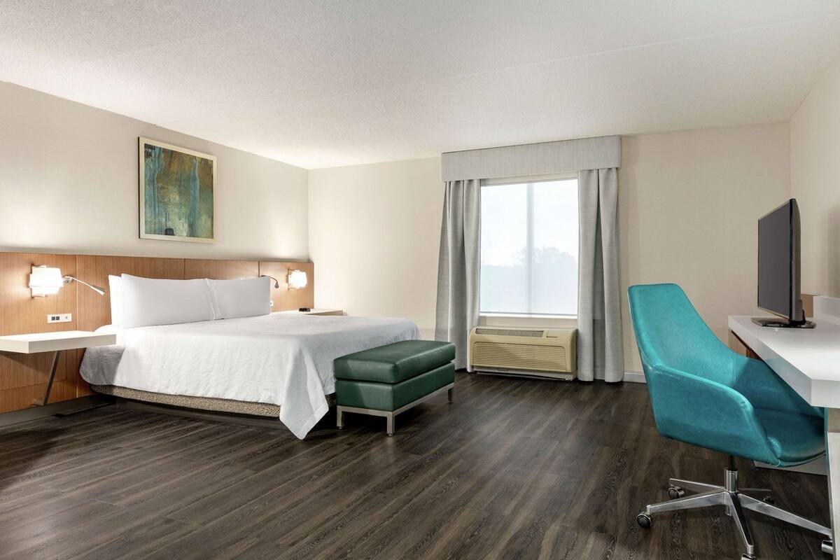 A hotel room with a bed, a desk with a chair, a TV, a window with curtains, and a painting on the wall.