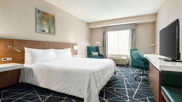 A hotel room with a large bed, a desk with a flat-screen TV, a teal chair, and a large window with curtains.