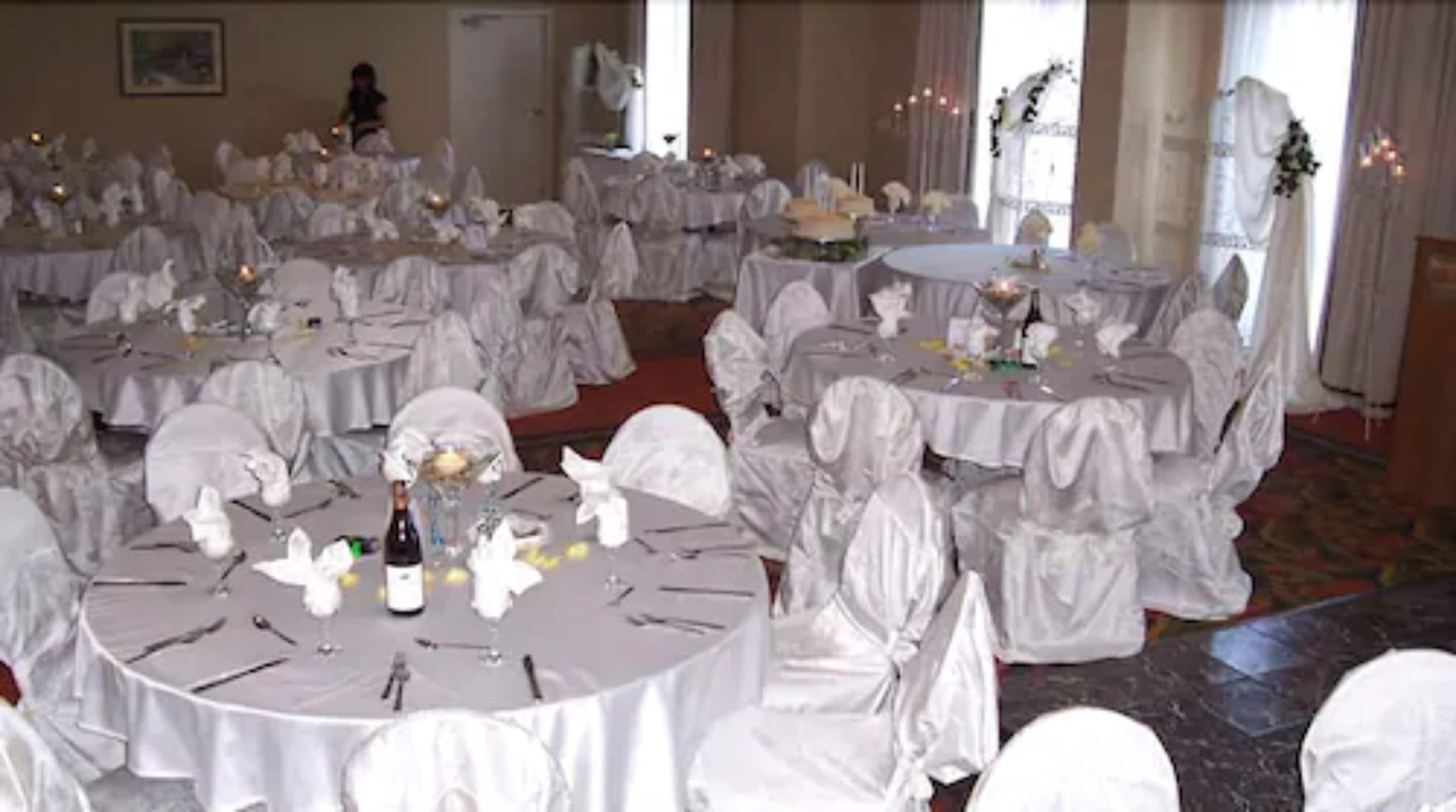 A banquet hall with round tables covered in white tablecloths, each set with plates, cutlery, and a bottle of wine.