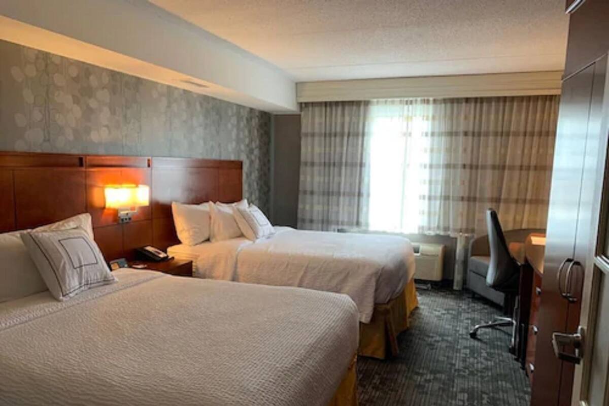 A hotel room with two beds, a wooden headboard, a nightstand with a lamp, a window with curtains, and a desk with a chair.