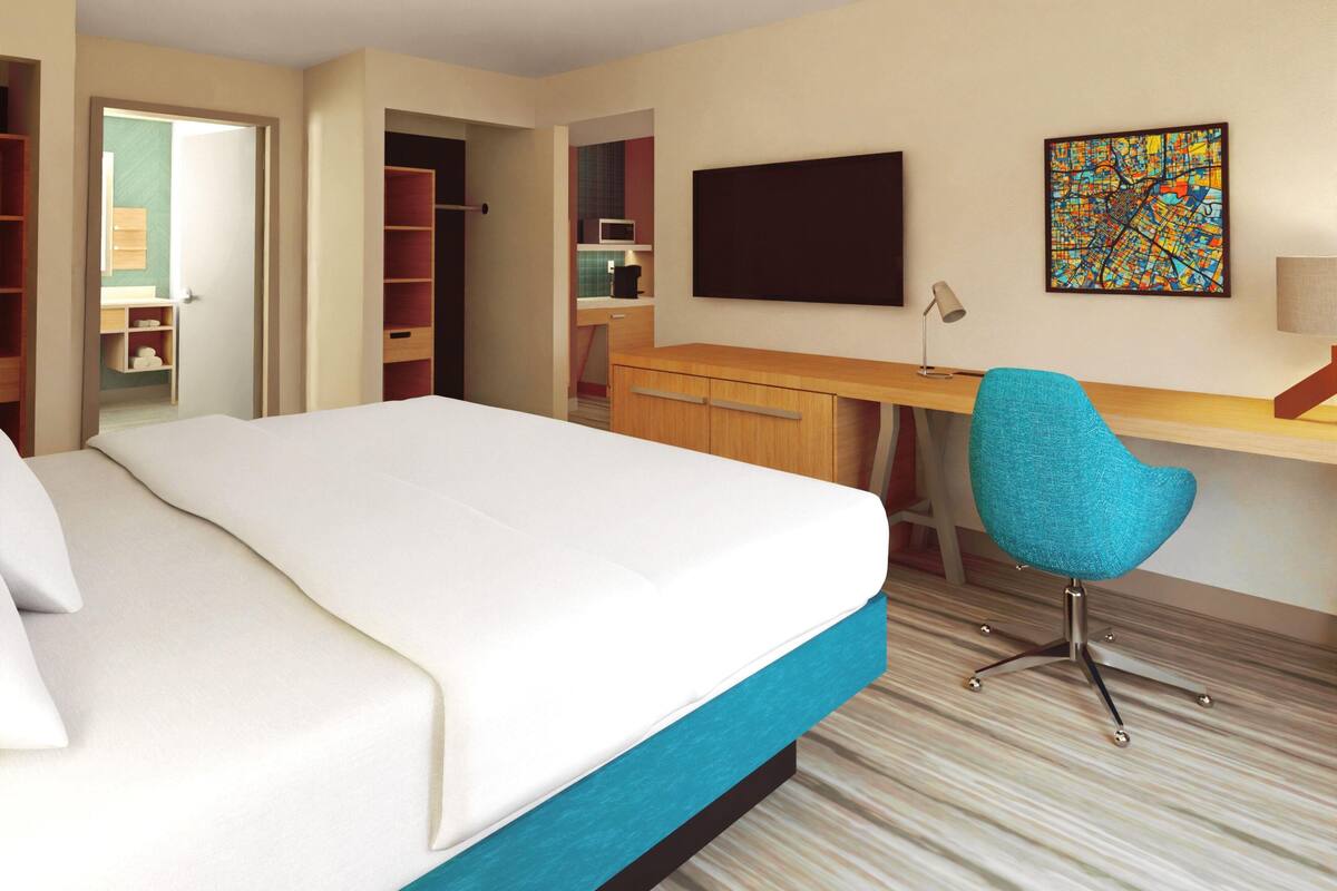 A hotel room with a bed, a desk, a chair, a television, and a colorful artwork on the wall.
