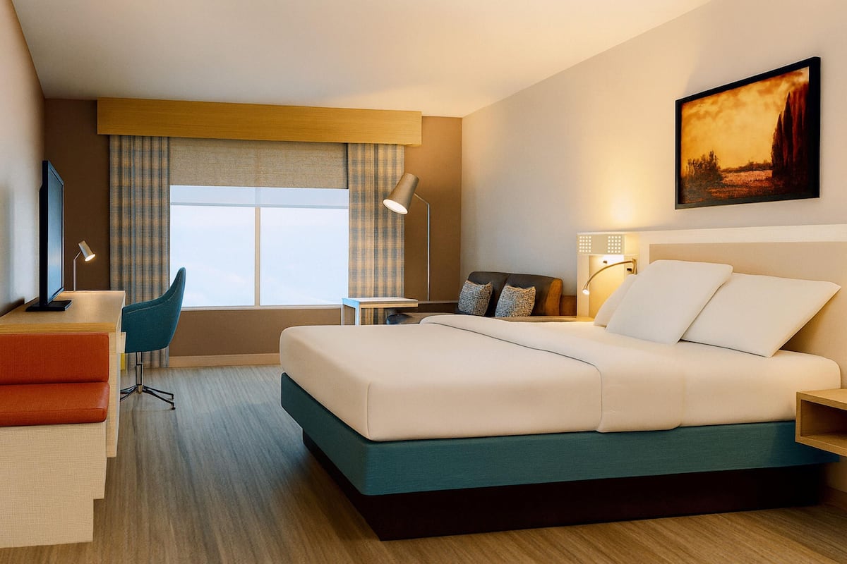 A hotel room with a bed, desk, chair, bedside table, lamp, and a painting on the wall.