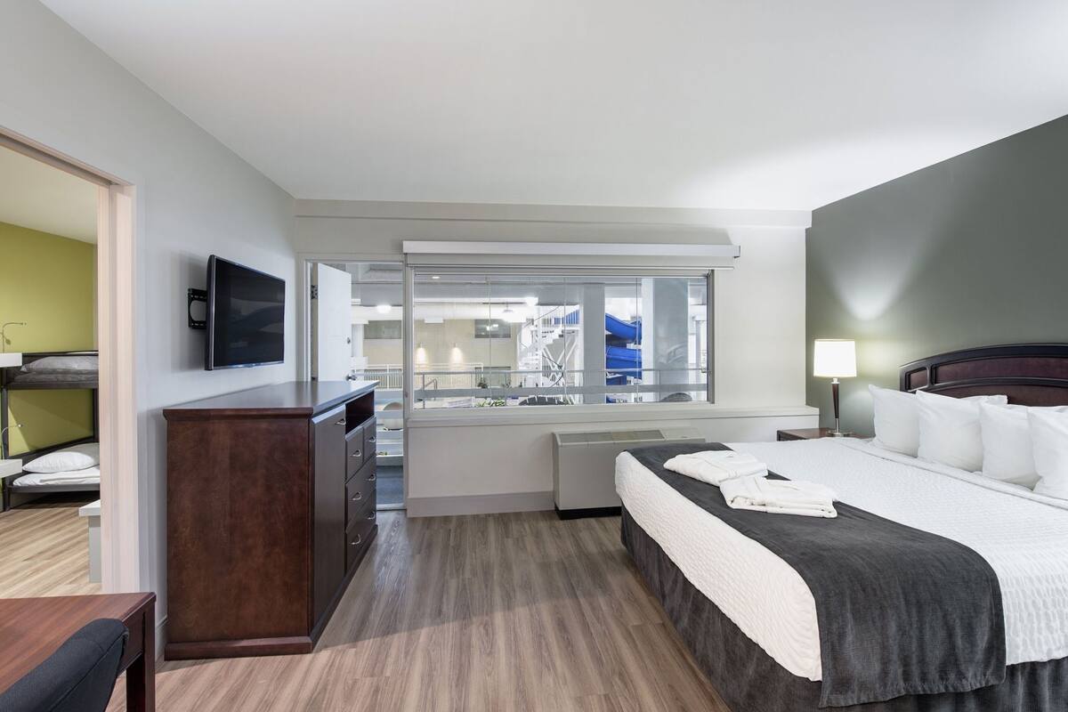 A hotel room with a bed, desk, TV, and a view of the city through a large window.