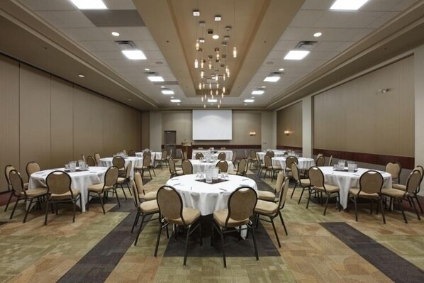 Une grande salle de conférence dotée de plusieurs tables rondes et de chaises, ainsi que d’un écran de projection et de luminaires au plafond.