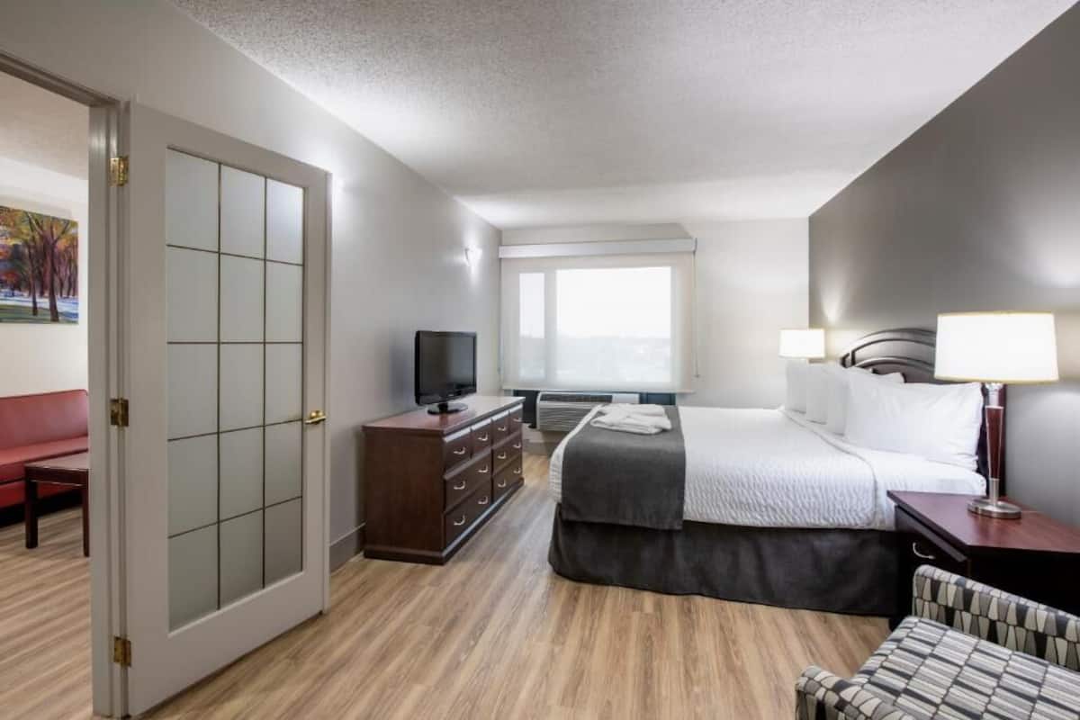 A hotel room with a bed, a TV, a dresser, a desk with a lamp, and a window with blinds.