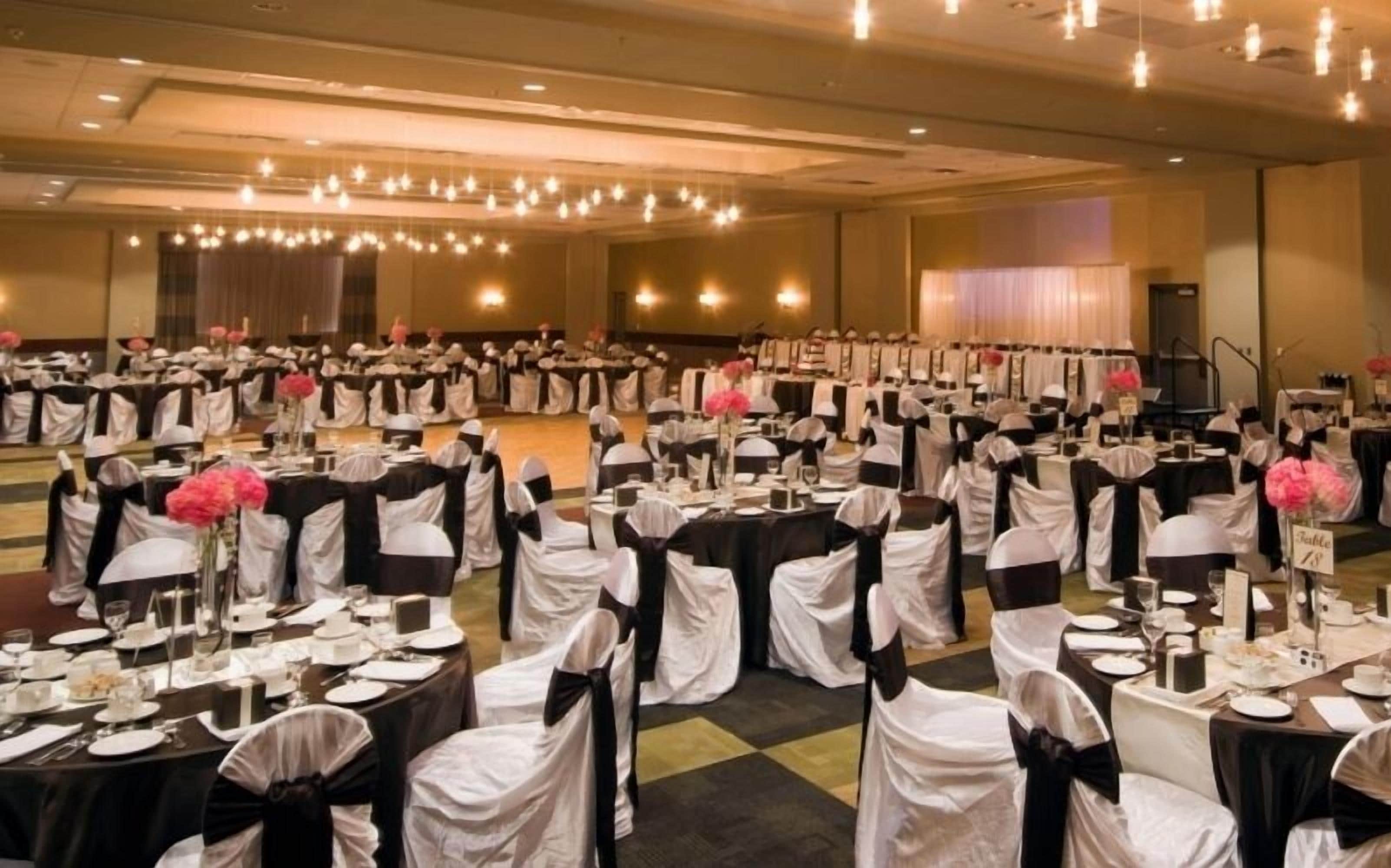 Un salón de banquetes con mesas dispuestas para un evento formal, con manteles blancos y negros, sillas con cintas negras y centros de mesa florales en tonos rosas.