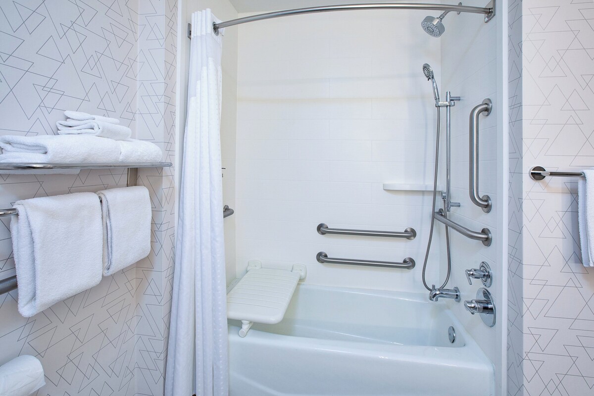 A modern bathroom with a walk-in shower, grab bars, a white bathtub, and neatly folded towels on a shelf.