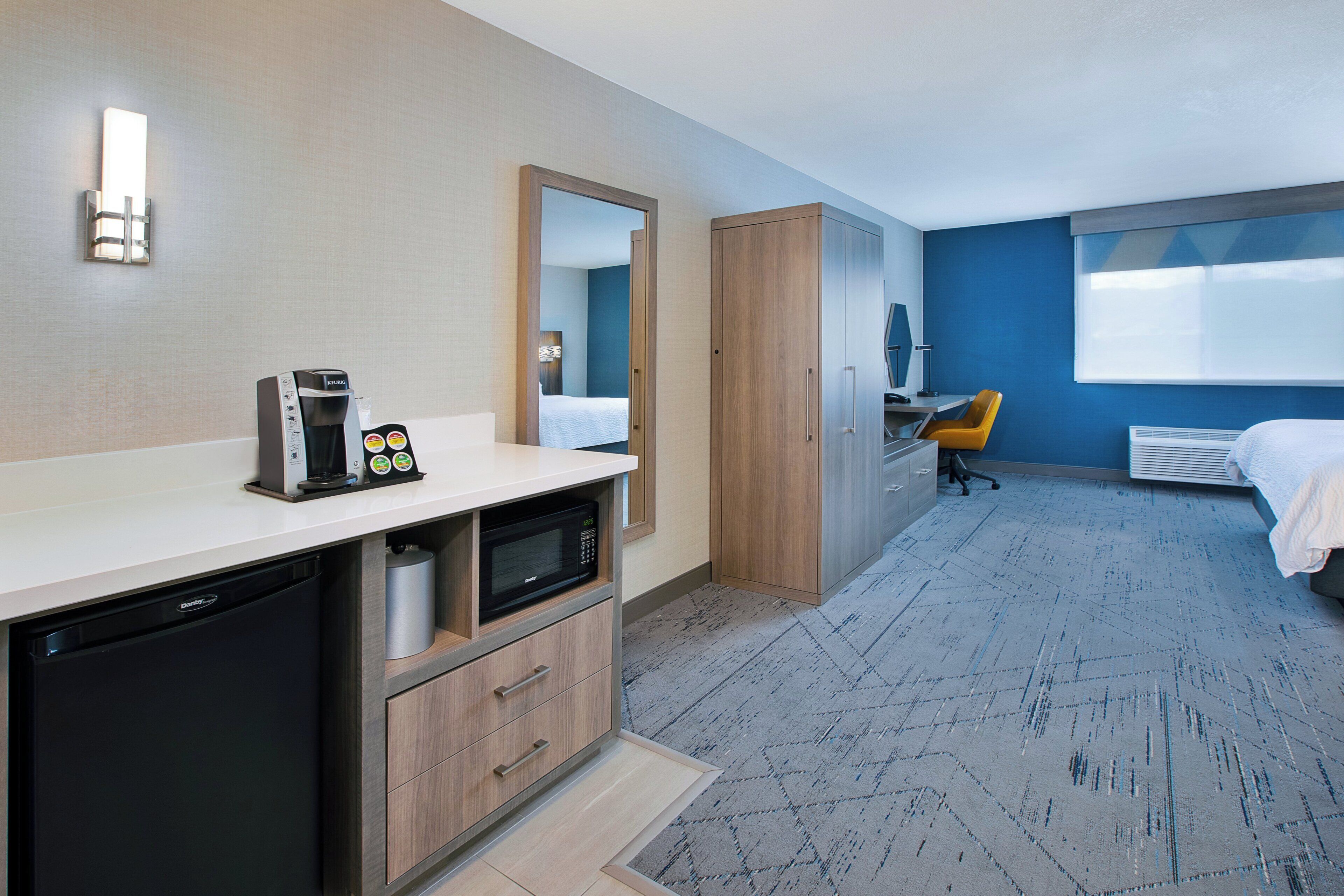 A hotel room with a kitchenette, a bed, a desk, and a large window.