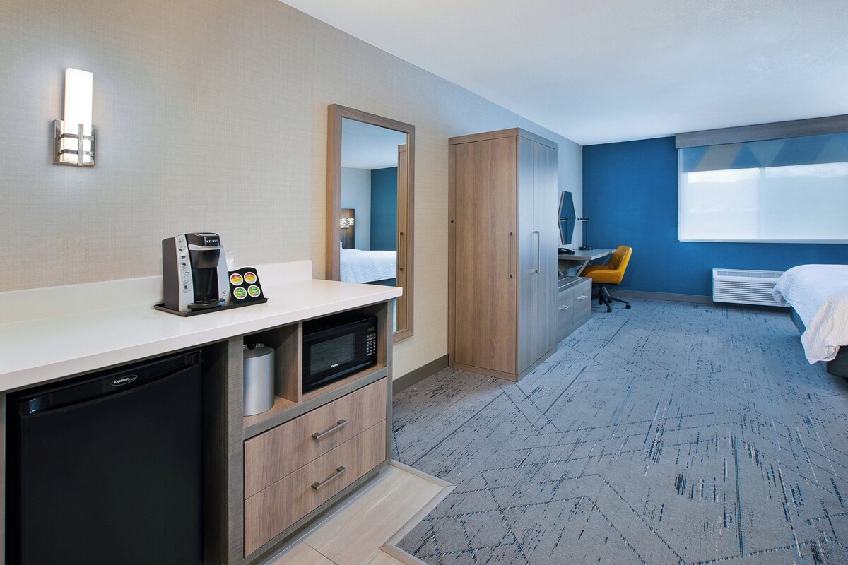 A hotel room with a kitchenette, a bed, a desk, and a large window.