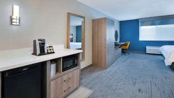 A hotel room with a kitchenette, a bed, a desk, and a large window.