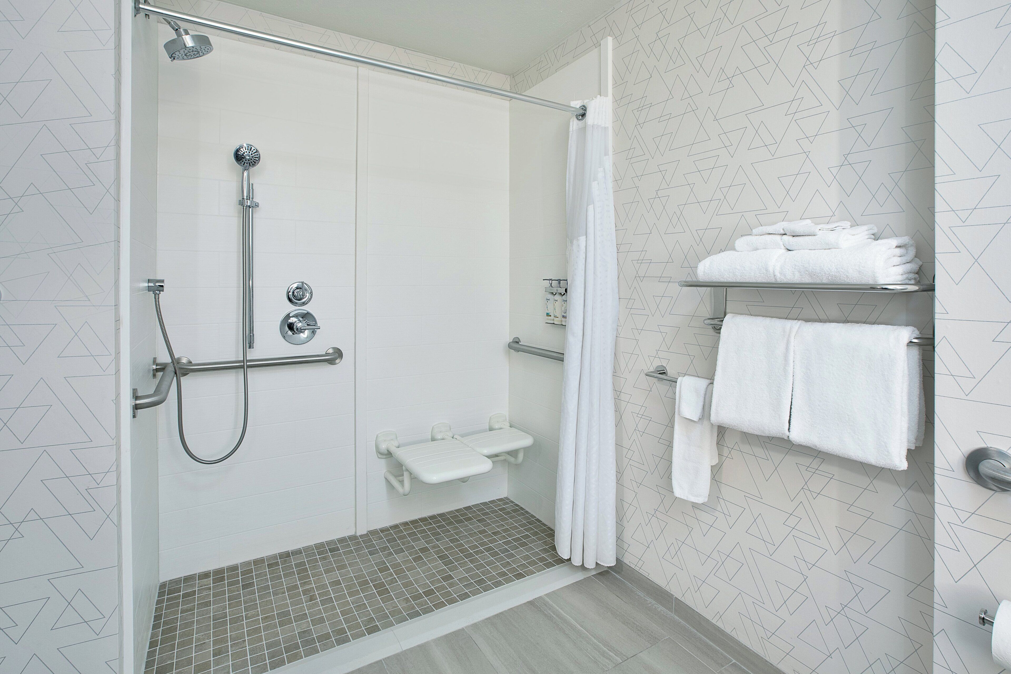 A modern shower area with a glass door, a handheld showerhead, a handheld shower attachment, a grab bar, a shelf with towels, and a tiled floor.