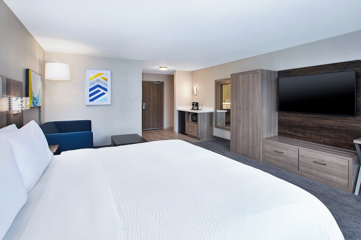 A hotel room with a large bed, a flat-screen TV, a desk with a chair, and a small kitchenette.