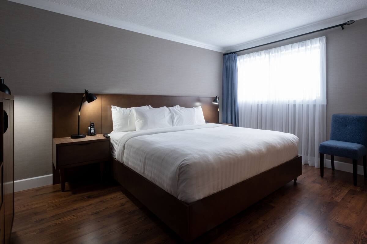 Ett modernt hotellrum med en stor säng, ett nattduksbord med en lampa, en blå stol och ett fönster med tunna, genomskinliga gardiner.