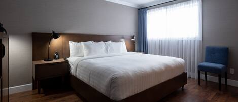 A modern hotel room with a large bed, a nightstand with a lamp, a blue chair, and a window with sheer curtains.