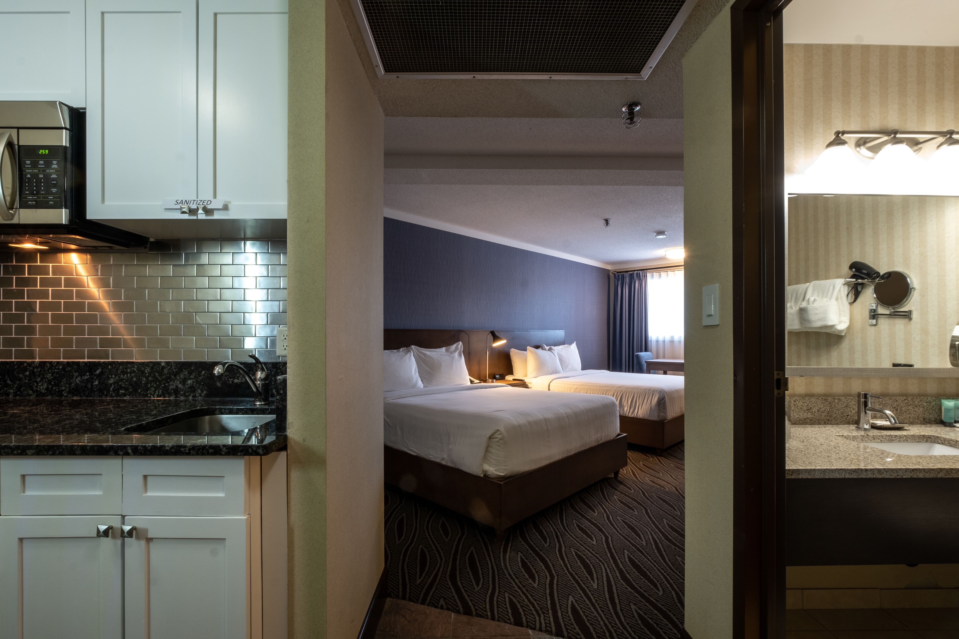 Une chambre d’hôtel avec une kitchenette, deux lits, une salle de bain avec un miroir et une moquette à motifs.