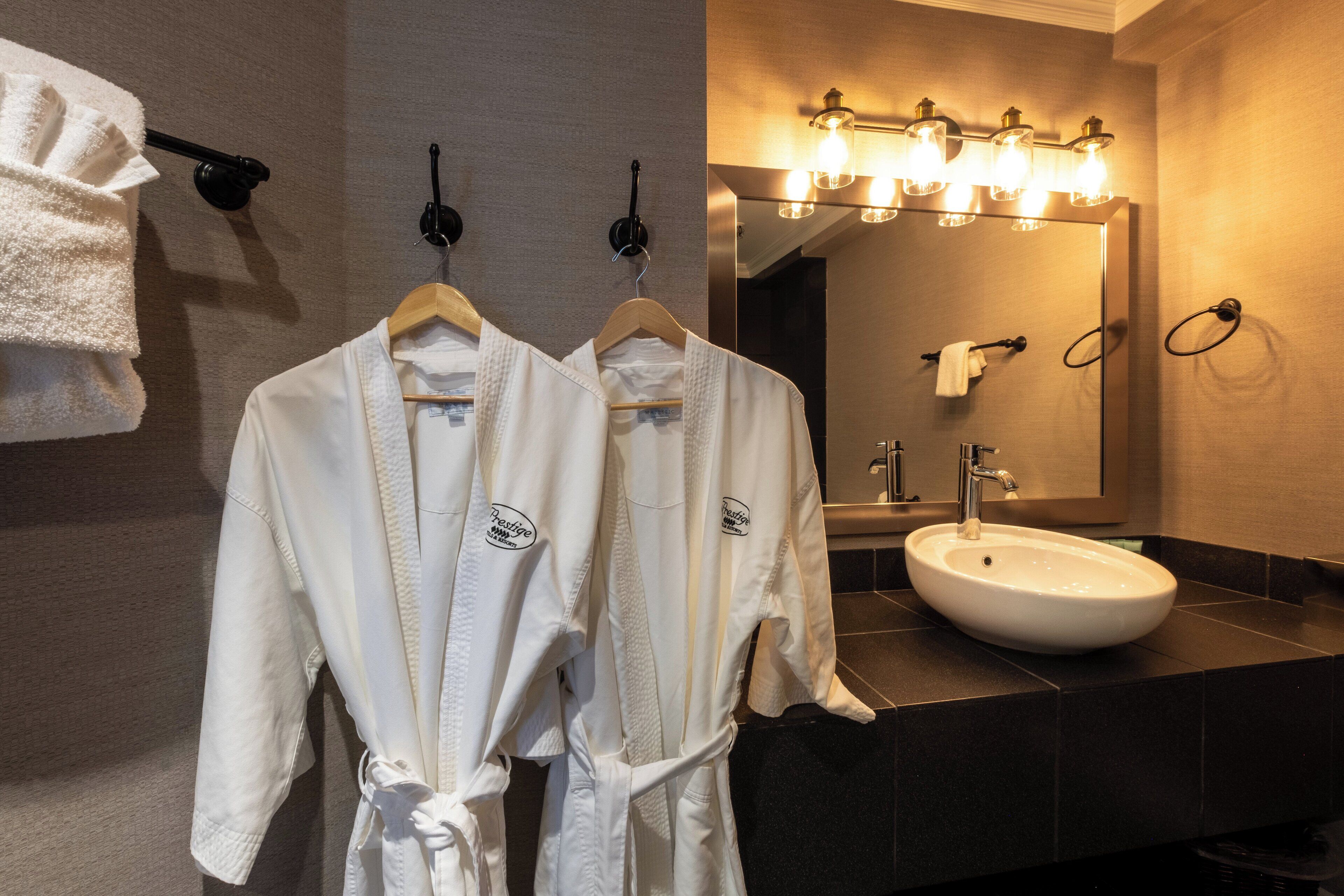 Une salle de bain avec deux peignoirs blancs suspendus à un porte-serviettes, un lavabo et un miroir avec des lumières au-dessus.