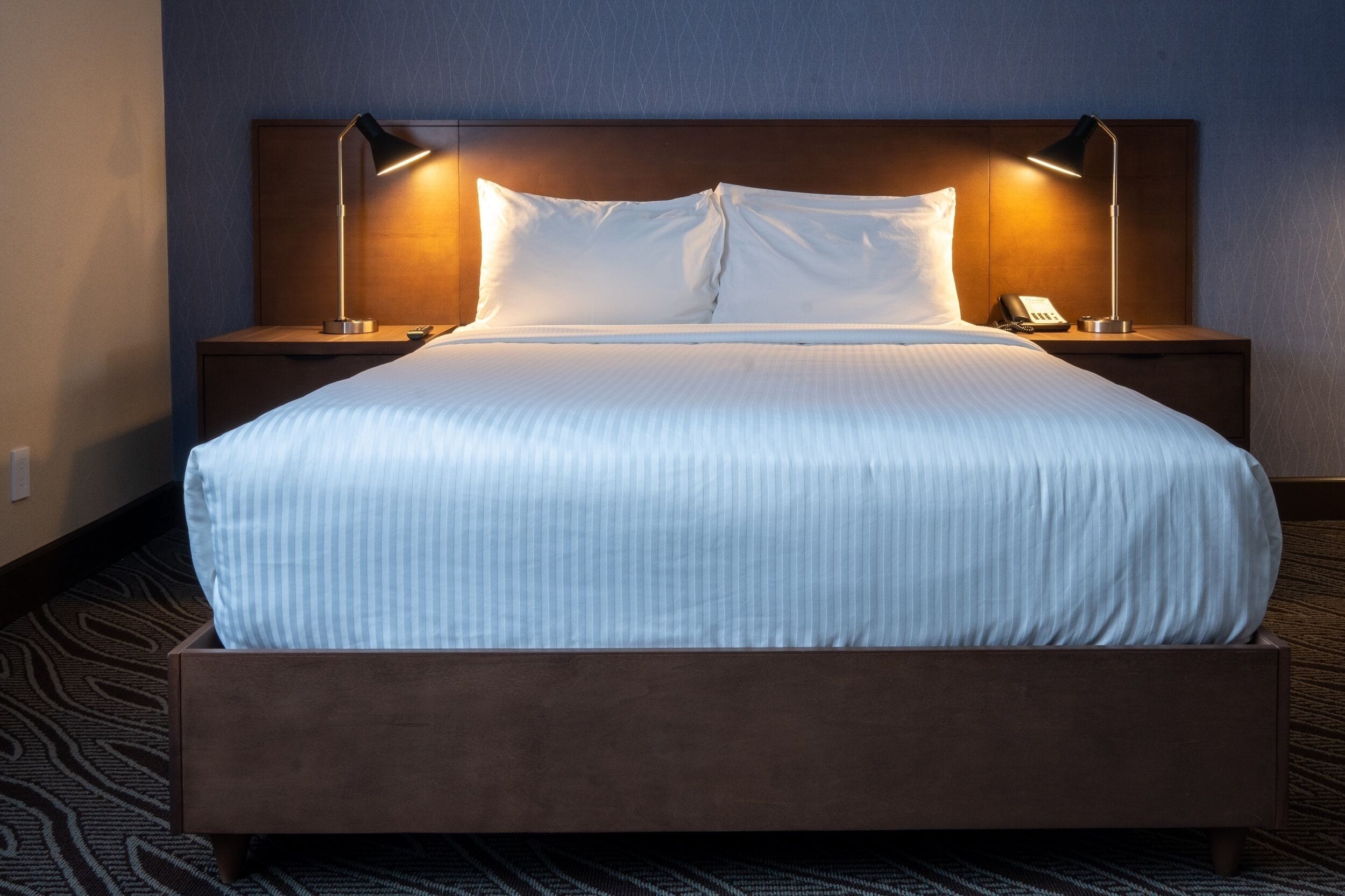 Una cama bien hecha con tablero de madera, dos lámparas de noche y un teléfono en la mesita.