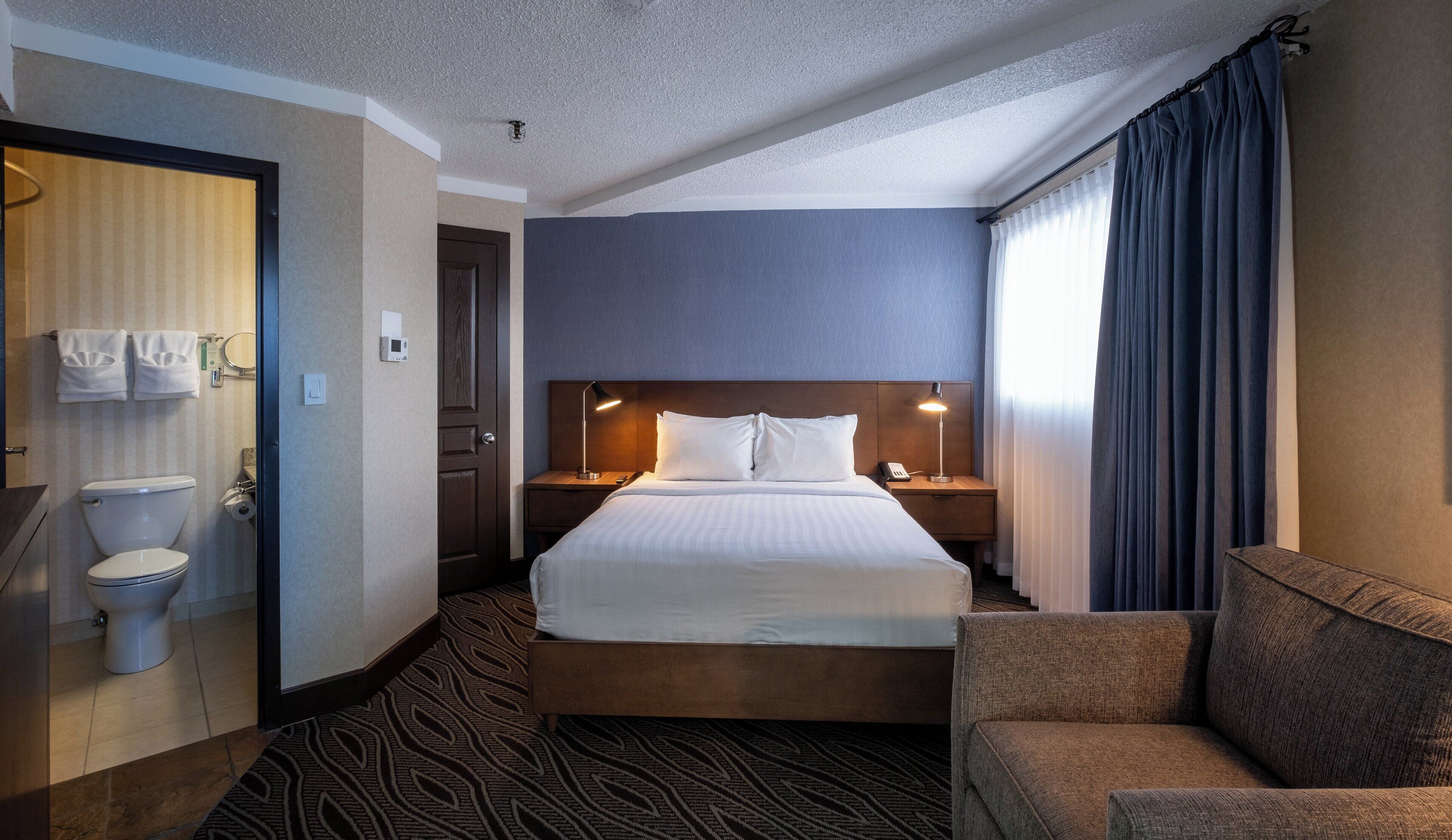 A hotel room with a bed, a bedside table with a lamp, a chair, a bathroom with a toilet and towel rack.