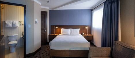 A hotel room with a bed, a bedside table with a lamp, a chair, a bathroom with a toilet and towel rack.