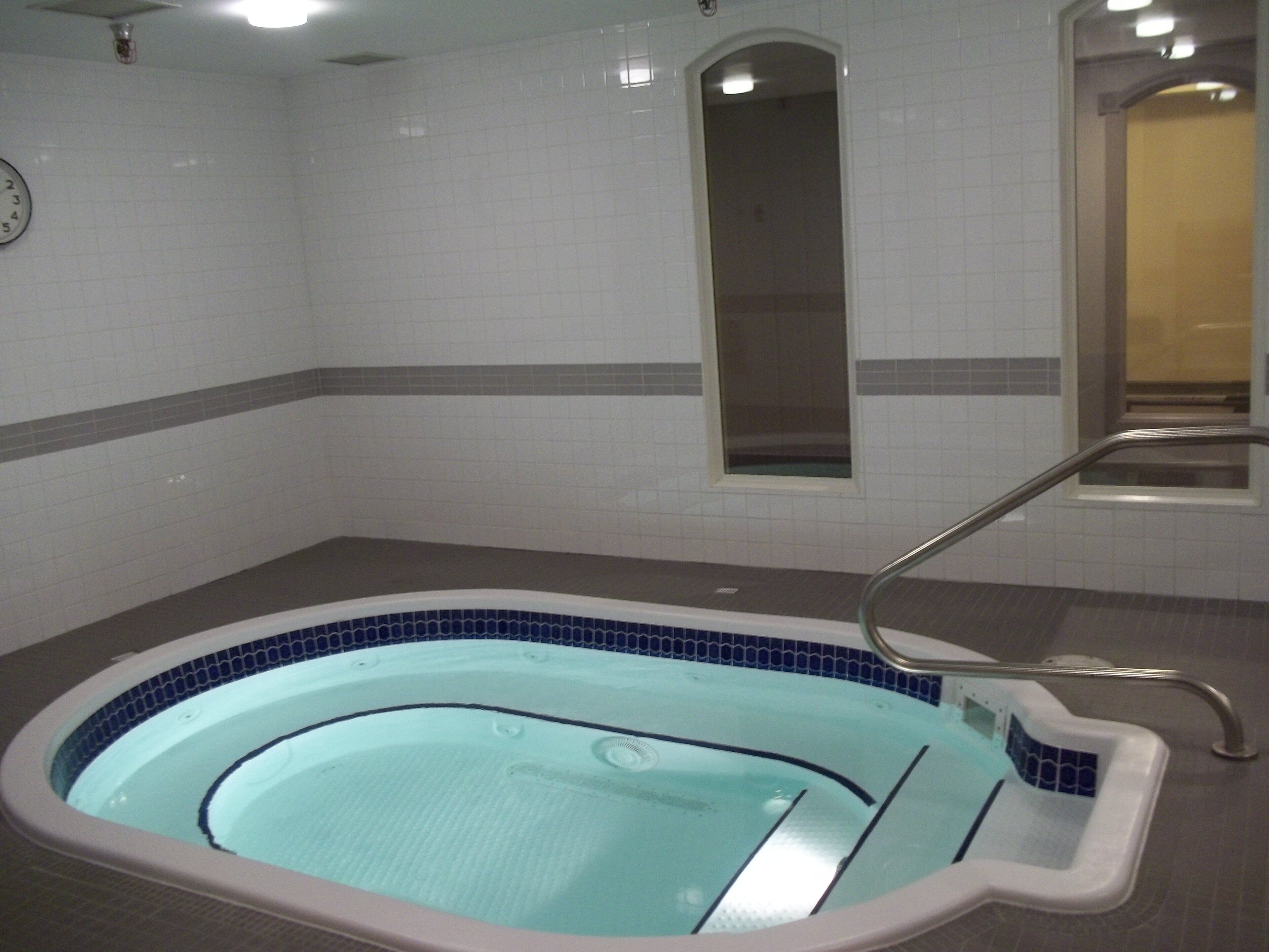 A small indoor swimming pool with a tiled border, a metal ladder, and a wall-mounted clock.