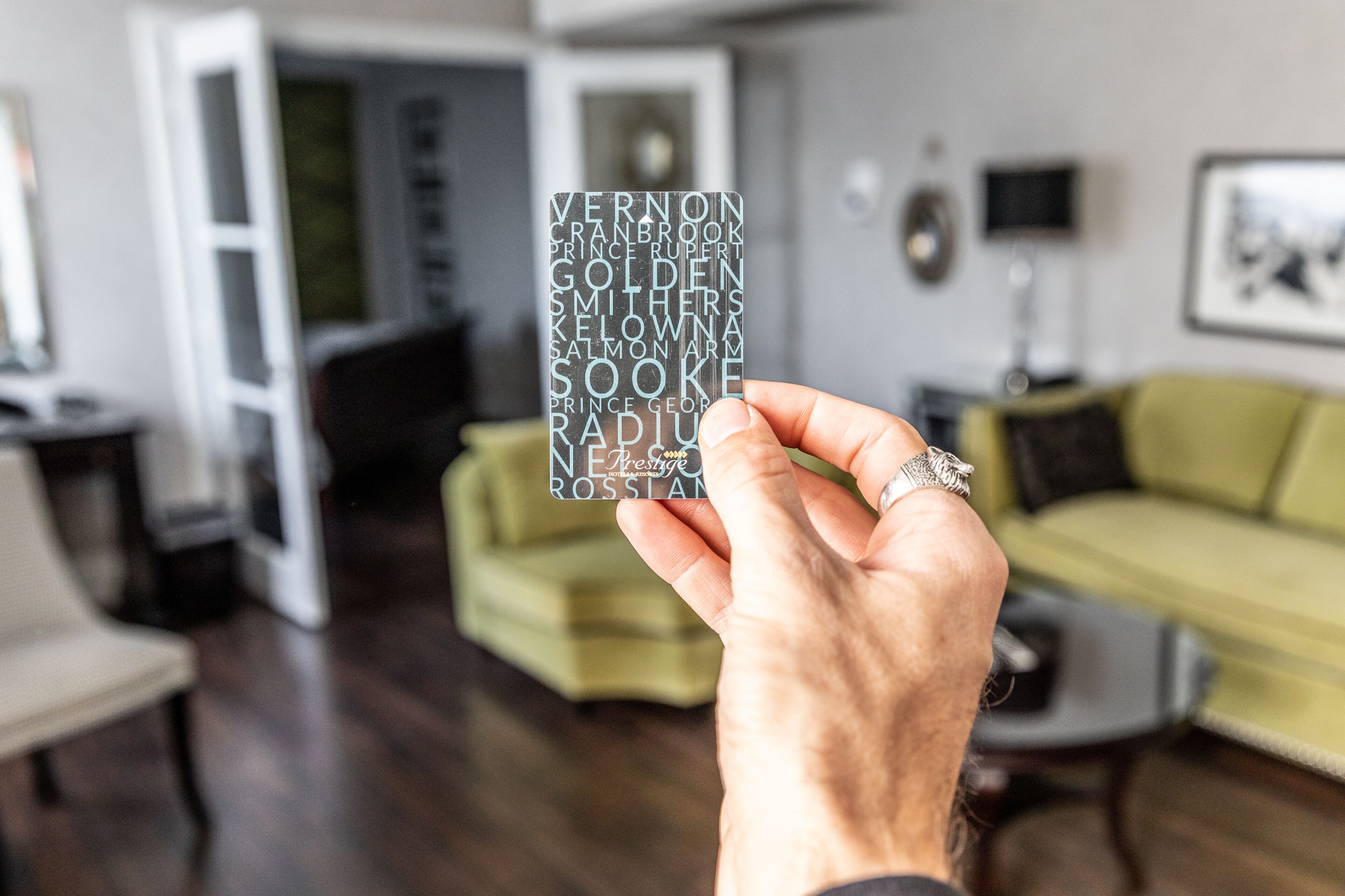A hand holding a card with names, in a living room with a green sofa, a coffee table, and a TV.